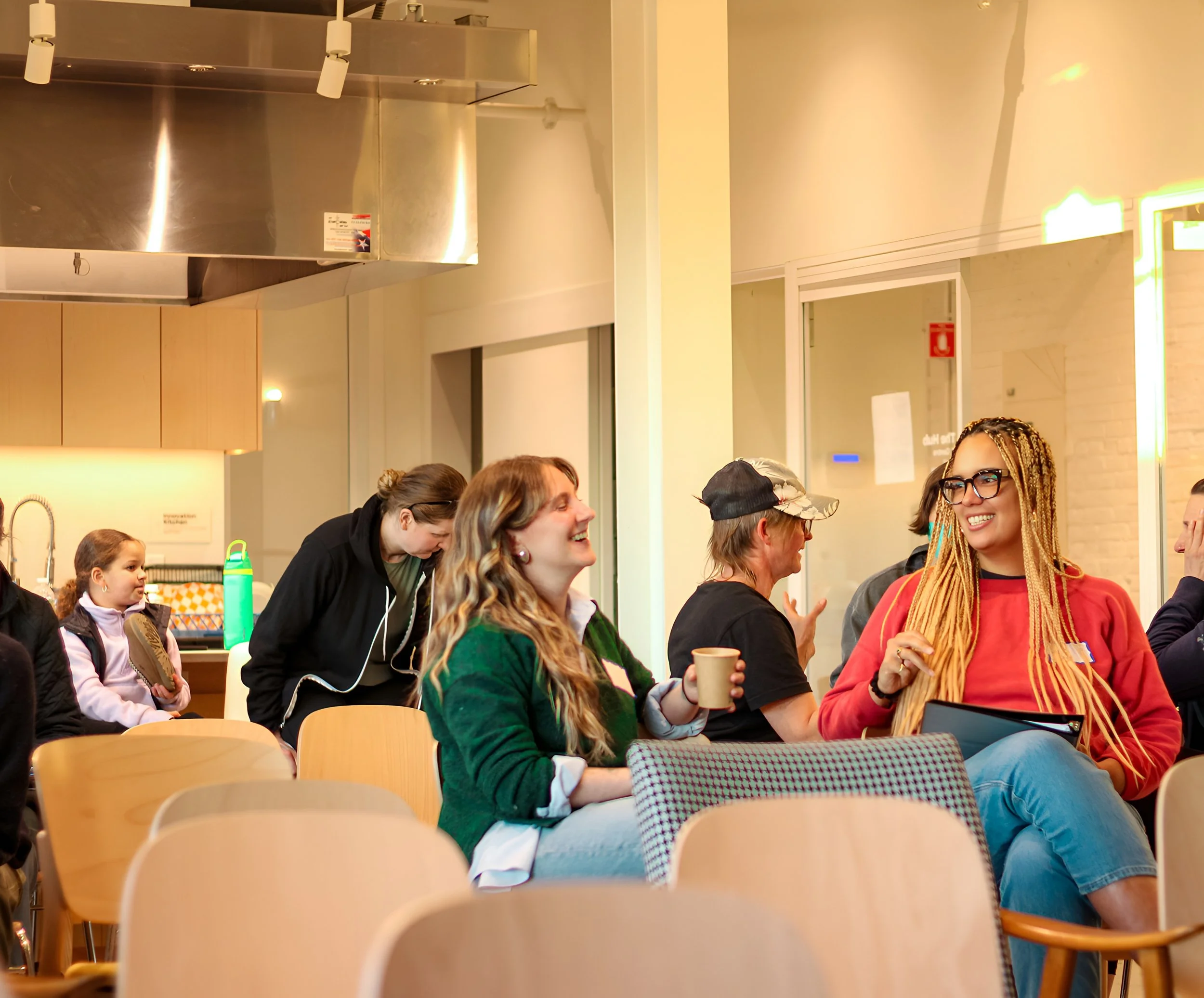 People sitting and standing in a meeting room, engaging in conversation and smiling, with casual attire and a cafeteria-like setting.