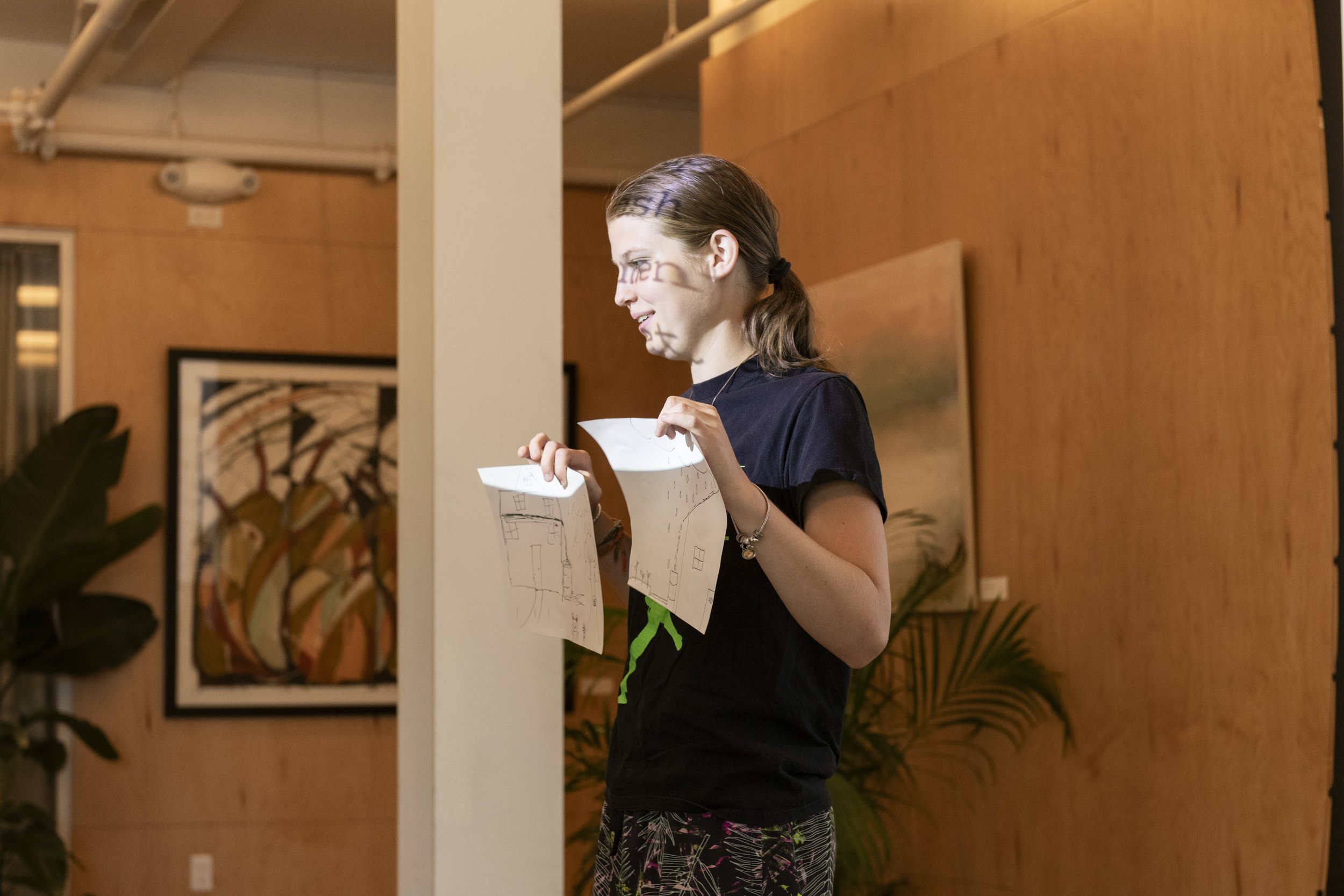 A young woman with brown hair tied back, standing indoors in a room with wooden walls, holding and looking at several sheets of paper with sketches on them, possibly diagrams or floor plans, during an art exhibition or presentation.