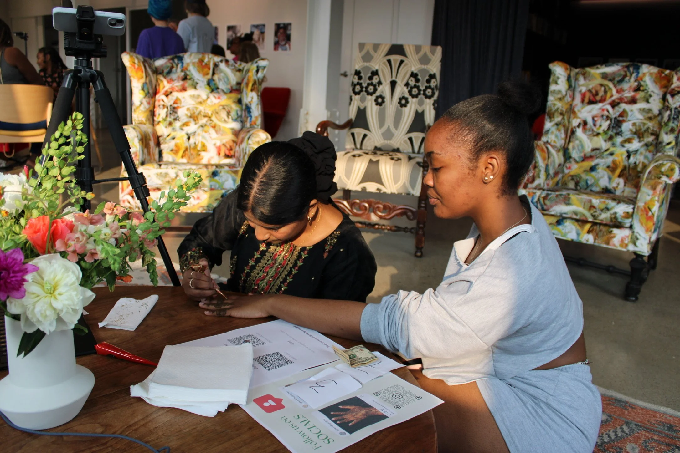 A woman with a ponytail gets a henna tattoo on her hand by another woman at a wooden table. The table has papers, a red pen, tissues, a QR code printout, and a bouquet of multicolored flowers. In the background, there are floral-patterned armchairs and people standing and talking.