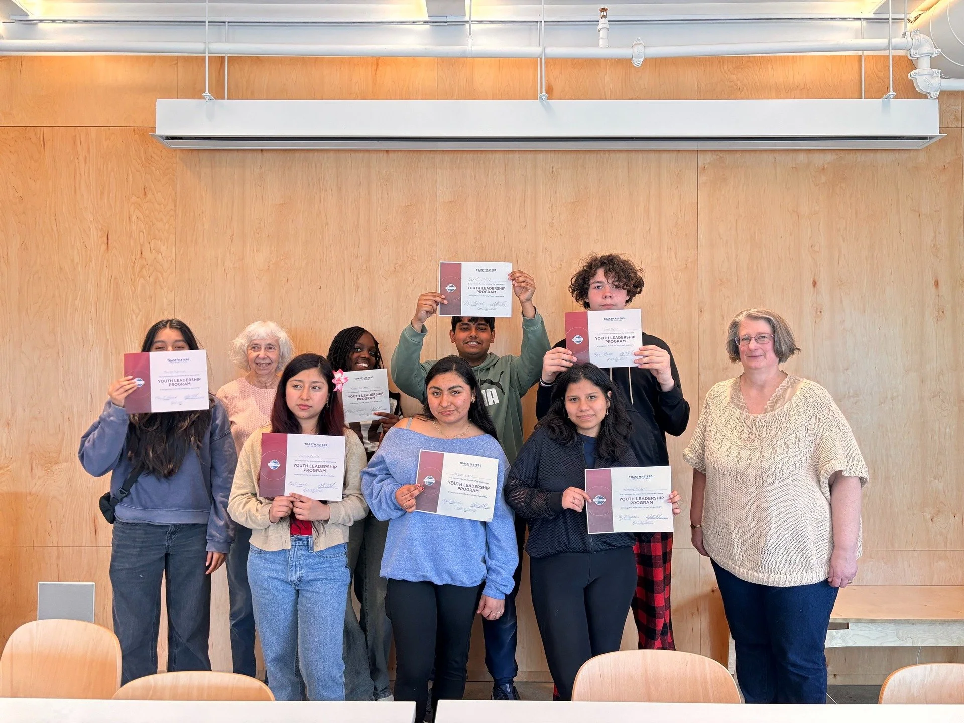 Group of ten people, mostly young, standing together holding certificates, in a wood-paneled room with a plain wall background, celebrating an achievement.