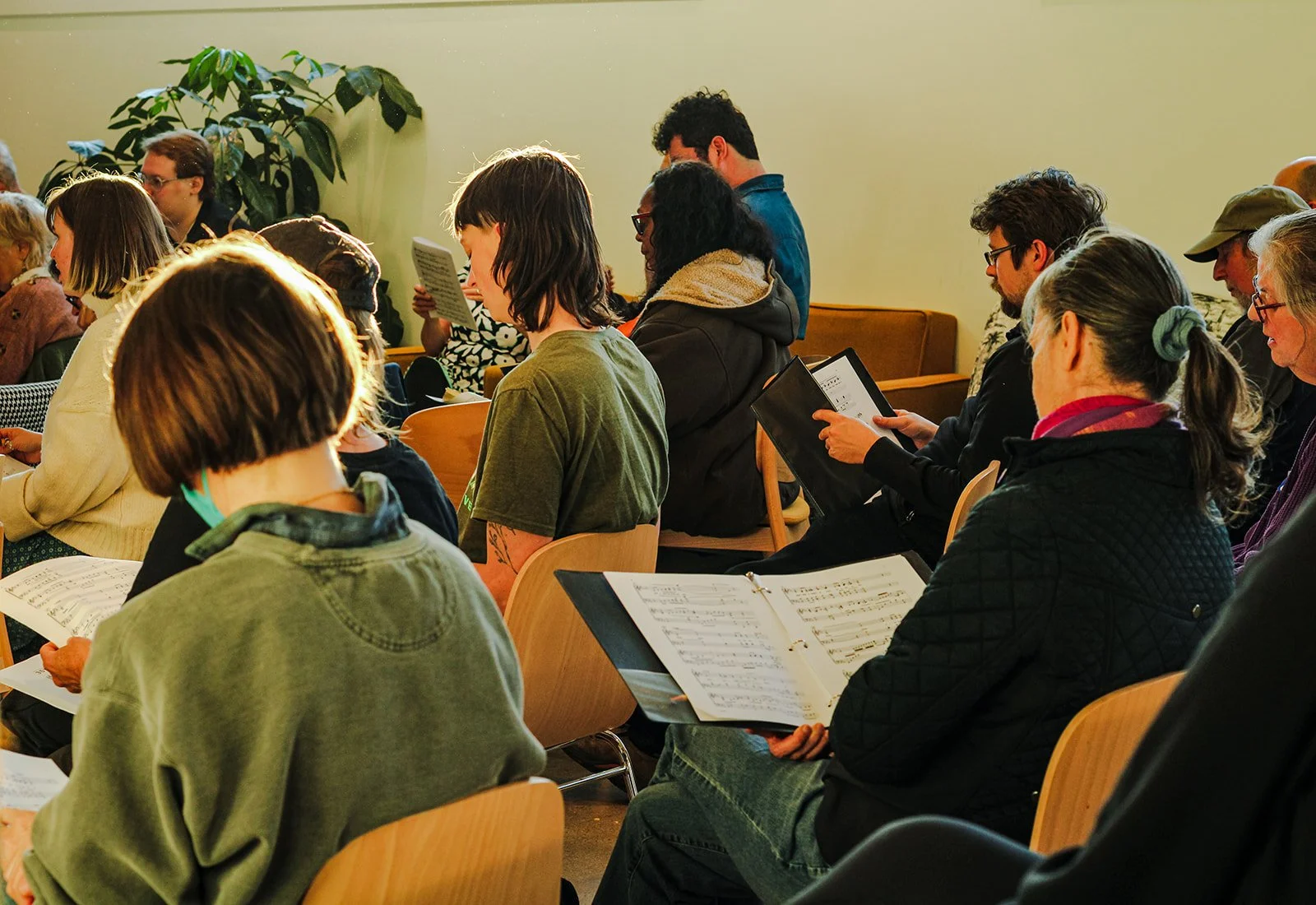 People attending a meeting or service, seated in rows holding sheets of music or programs, with some reading, in a room with green walls and a large potted plant.