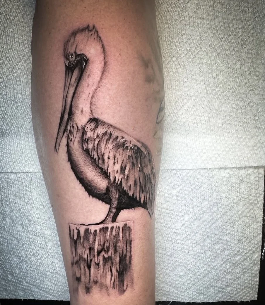 Black and gray tattoo of a pelican perched on a post on a person's thigh. black and grey realism tattoo pelican tattoo in Stuart, Sewall’s Point, Hutchinson Island, Jensen Beach, Palm City, Port St. Lucie, Fort pierce, treasure coast and surrounding 