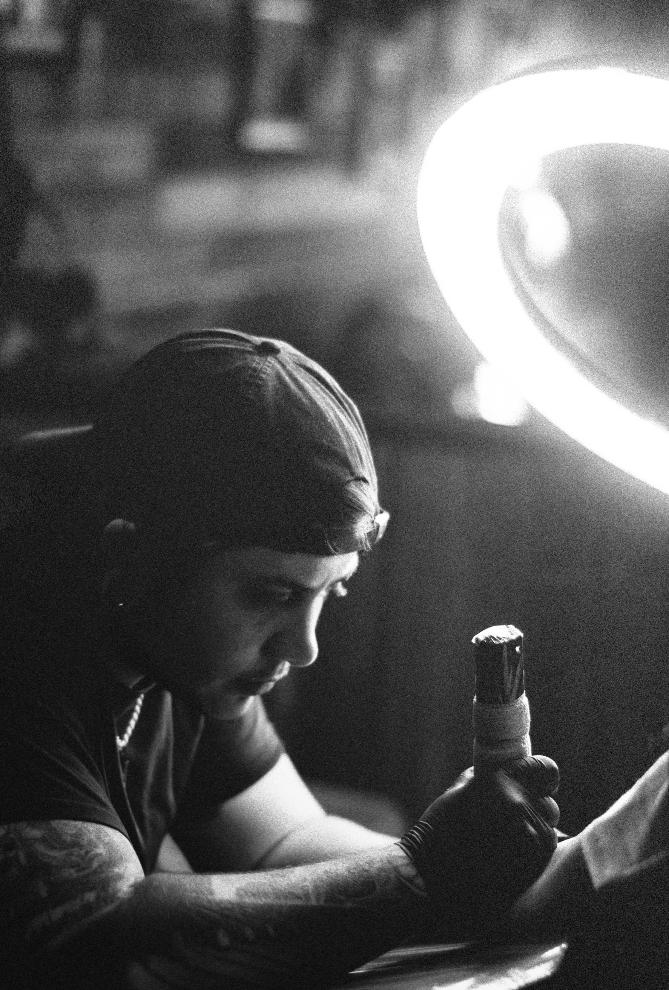 A man in a baseball cap and glove showing a tattooed arm, holding a tattoo machine in a dimly lit environment with a bright light nearby.
