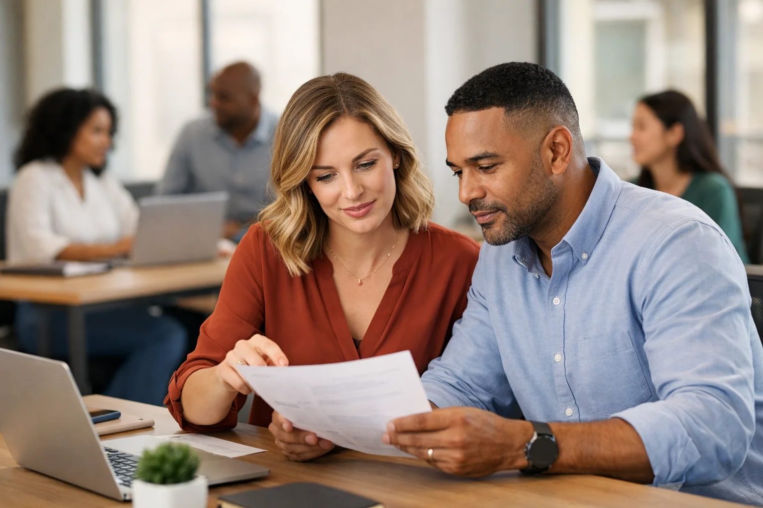 A man and woman sitting at a desk, looking at a document together, in a modern office setting with other colleagues working in the background.