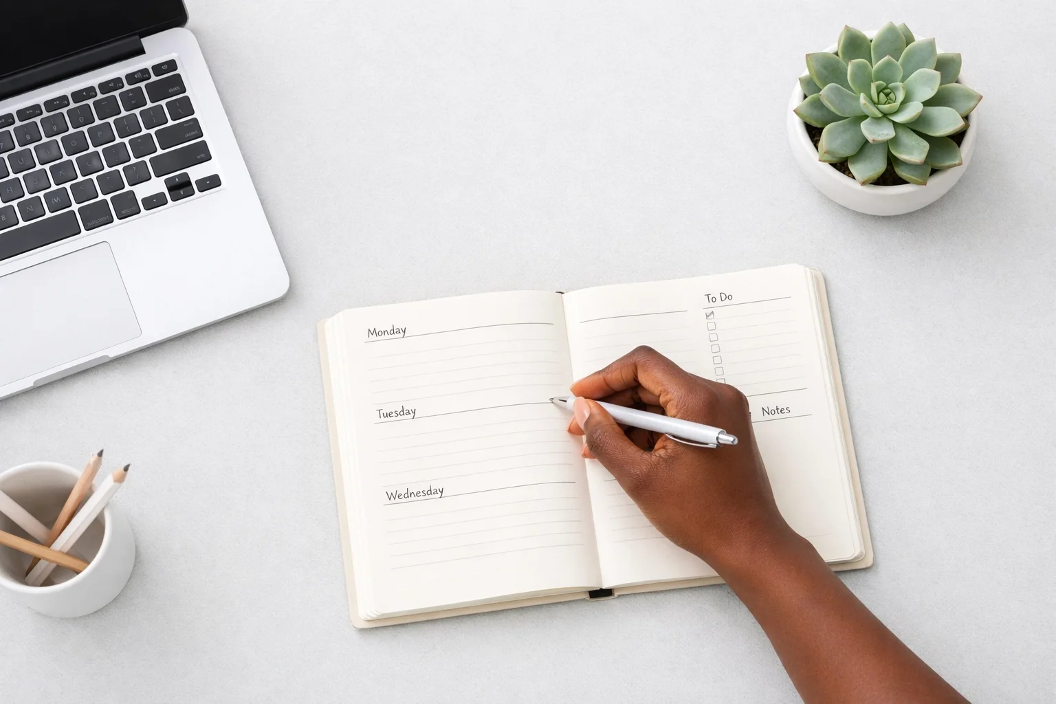 Open planner with days of the week, being written on by a hand, on a desk with a laptop, a small potted plant, a cup with pencils, and a notepad.