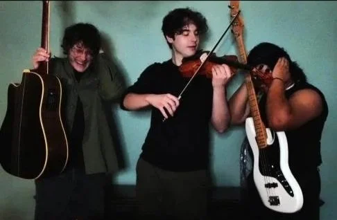Three young people with musical instruments against a teal wall, one holding an acoustic guitar, one playing a violin, and the third with a guitar covering their face.