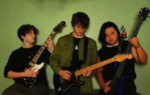 Three young men playing electric guitars against a green wall.