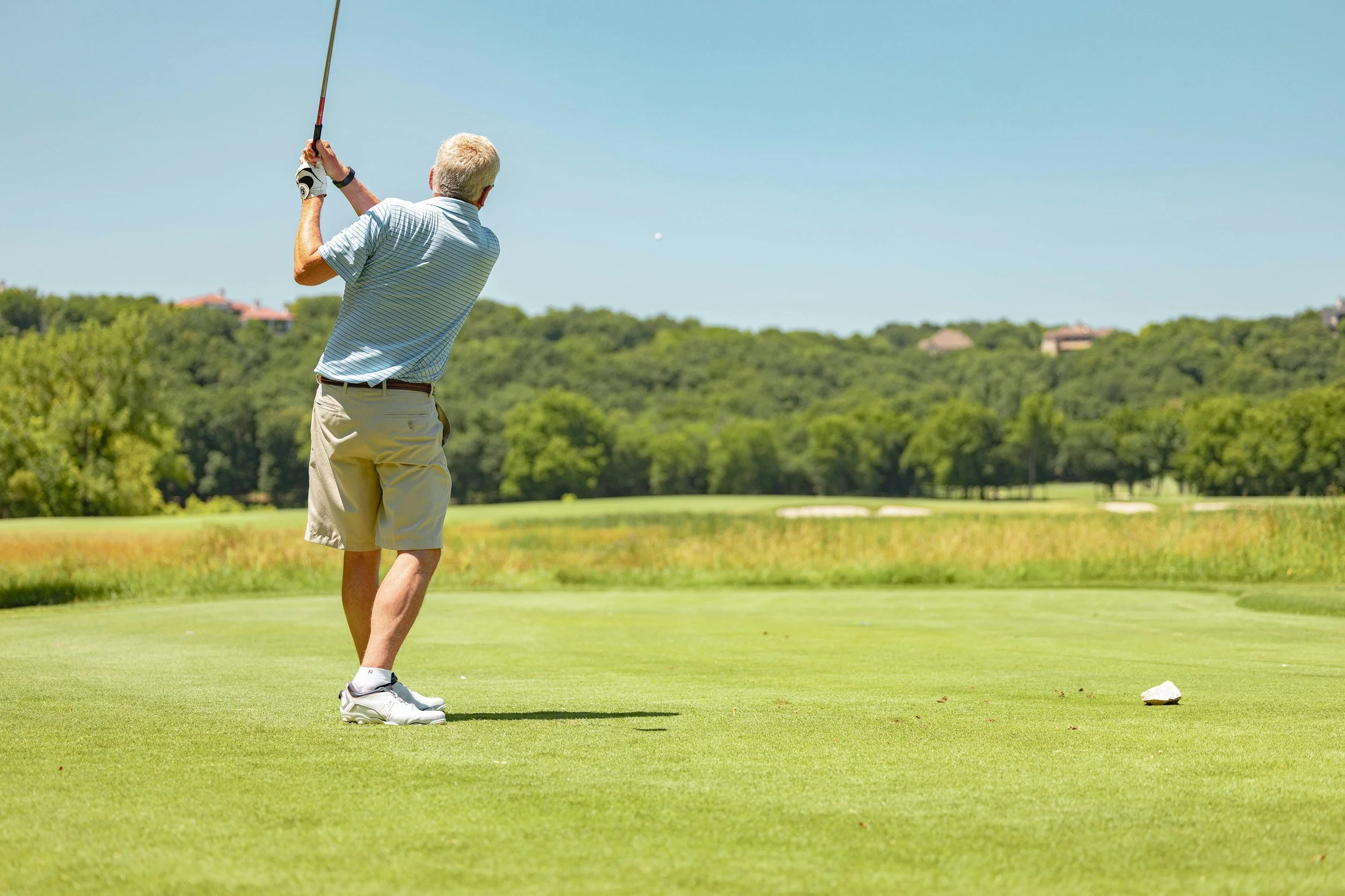 An older man with blond hair, wearing a striped polo shirt, beige shorts, and white sneakers, is swinging a golf club on a golf course with green grass and trees in the background.