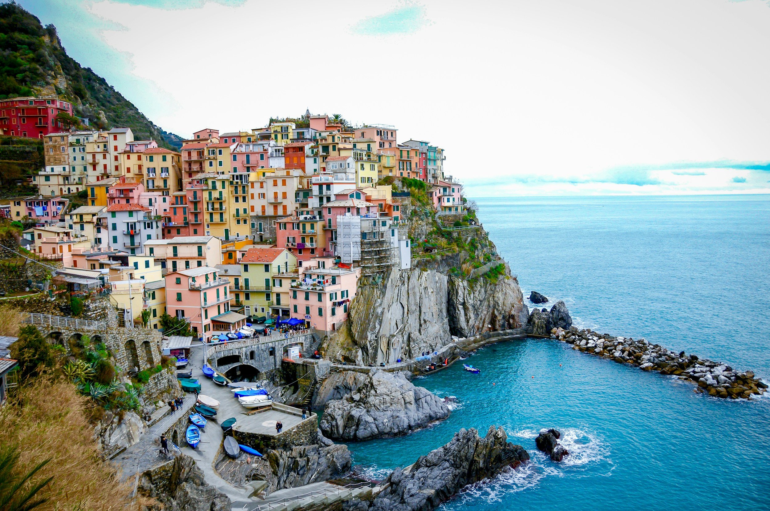 A hillside coastal town with multiple colorful houses overlooking the ocean, with boats anchored in the water at sunset.