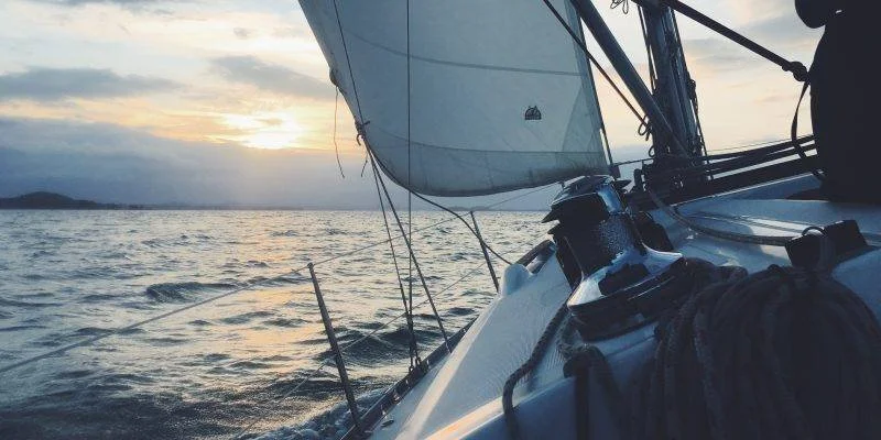 View from a sailboat at sunset, showing the boat's deck, sail, and the water with a distant shoreline.