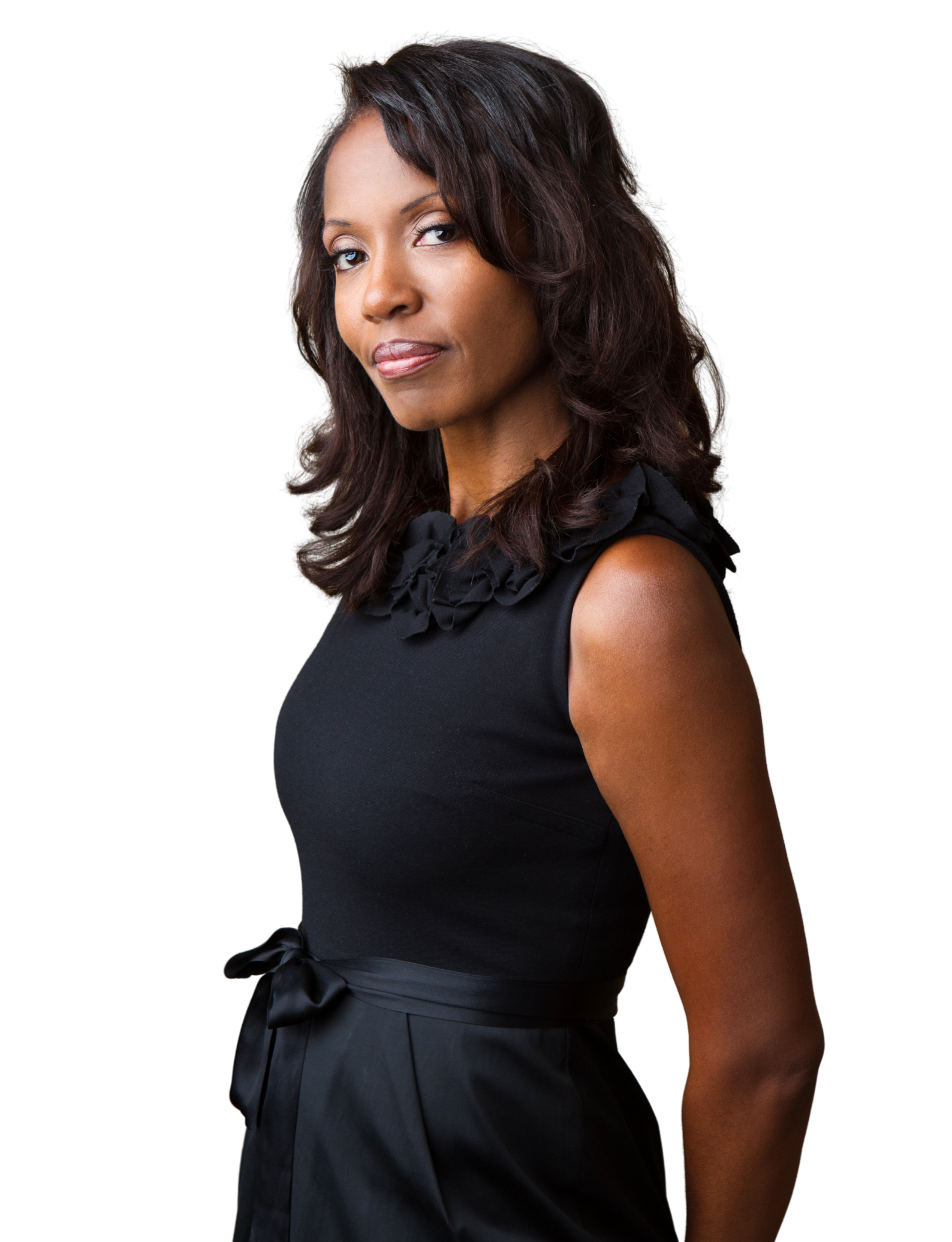 A woman with medium-length dark brown hair, wearing a sleeveless black dress with ruffle details around the neckline and a ribbon tied at the waist, looking confidently at the camera against a black background.