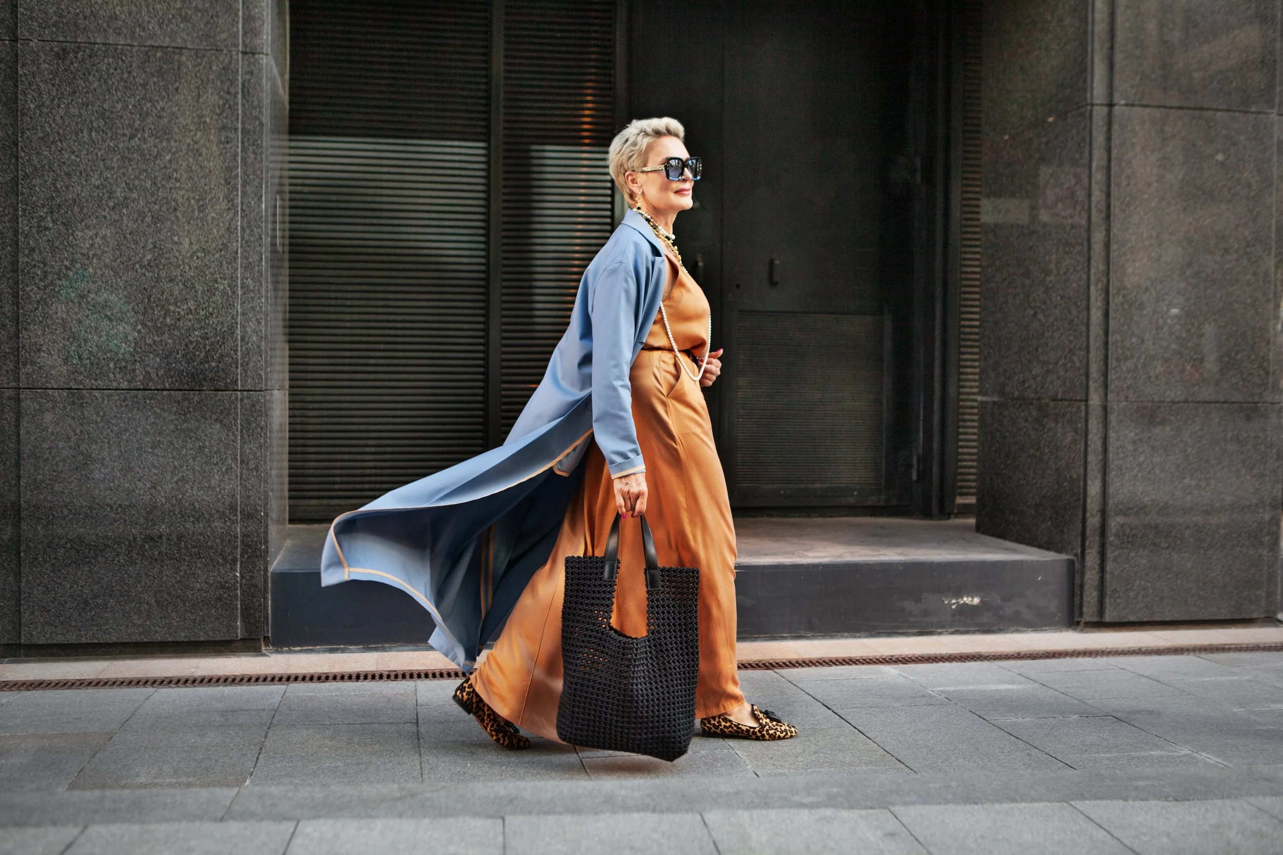 An elegant woman with short blonde hair wearing oversized sunglasses, a light blue trench coat, a bronze-colored dress, leopard-print flats, carrying a black woven tote bag, walking on a city sidewalk against a dark building background.