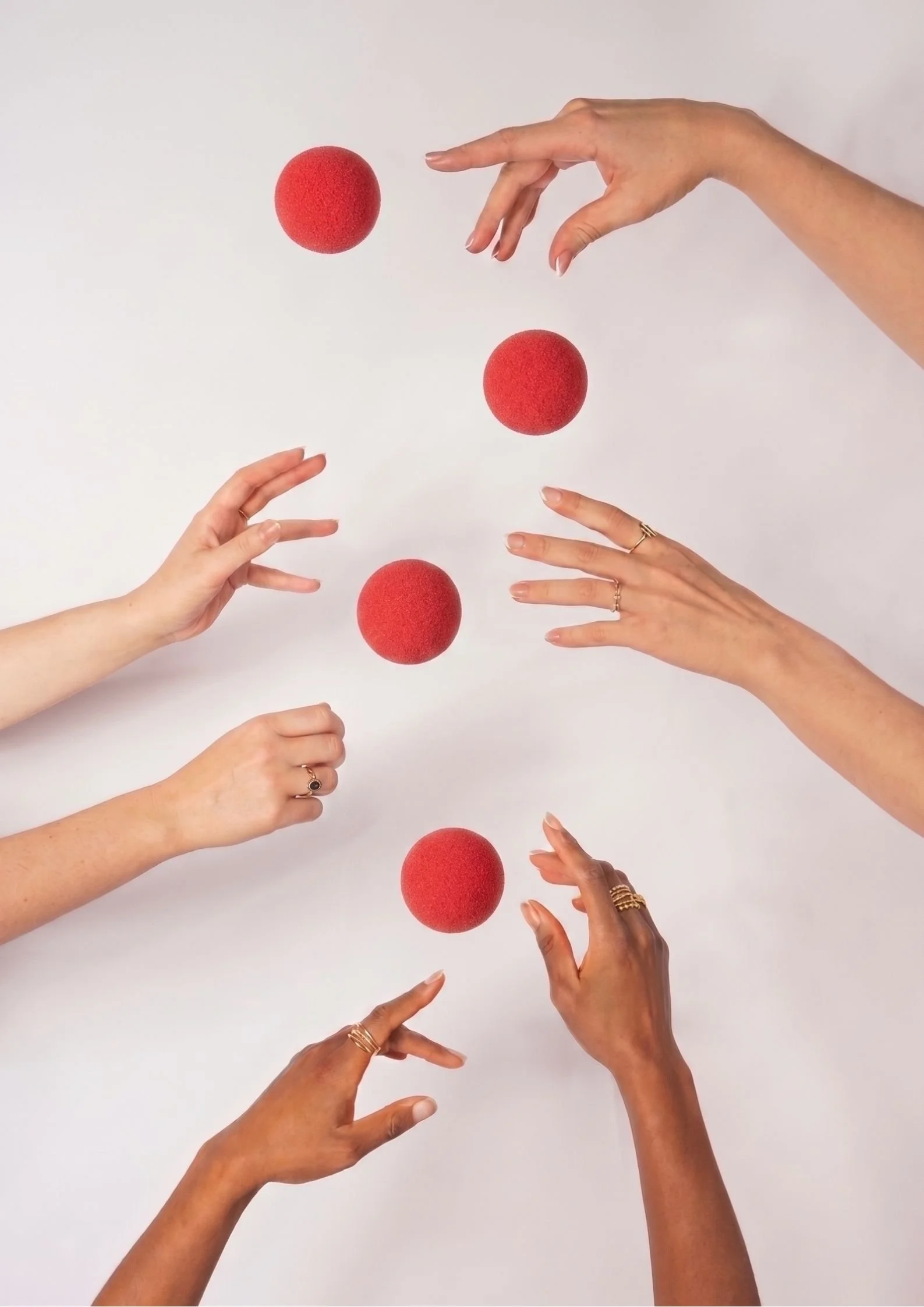 Multiple hands with various skin tones and gold rings are reaching towards four floating red clown noses against a pale background.