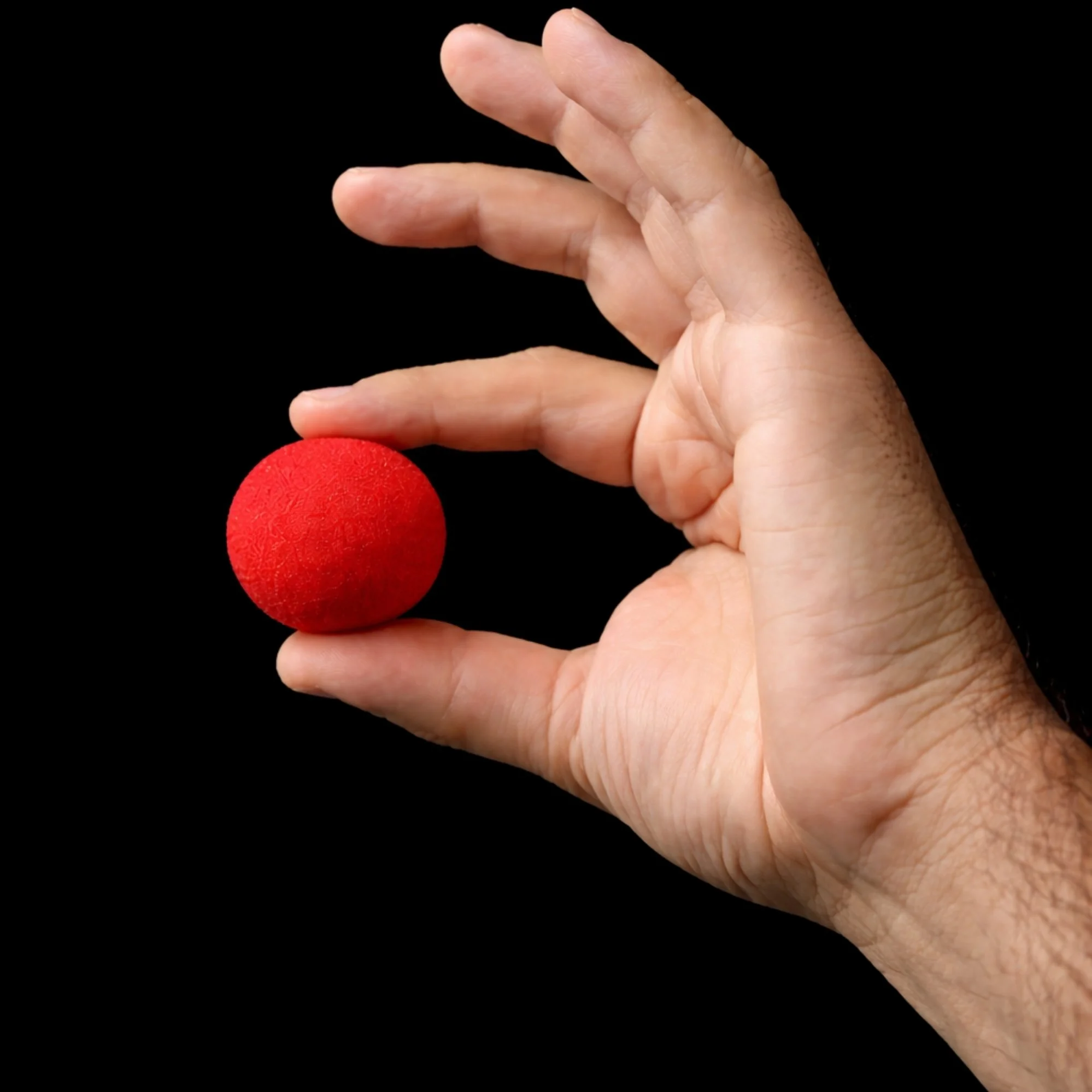 Close-up of a hand with fingers making a pinching gesture, holding a red clown nose against a black background.