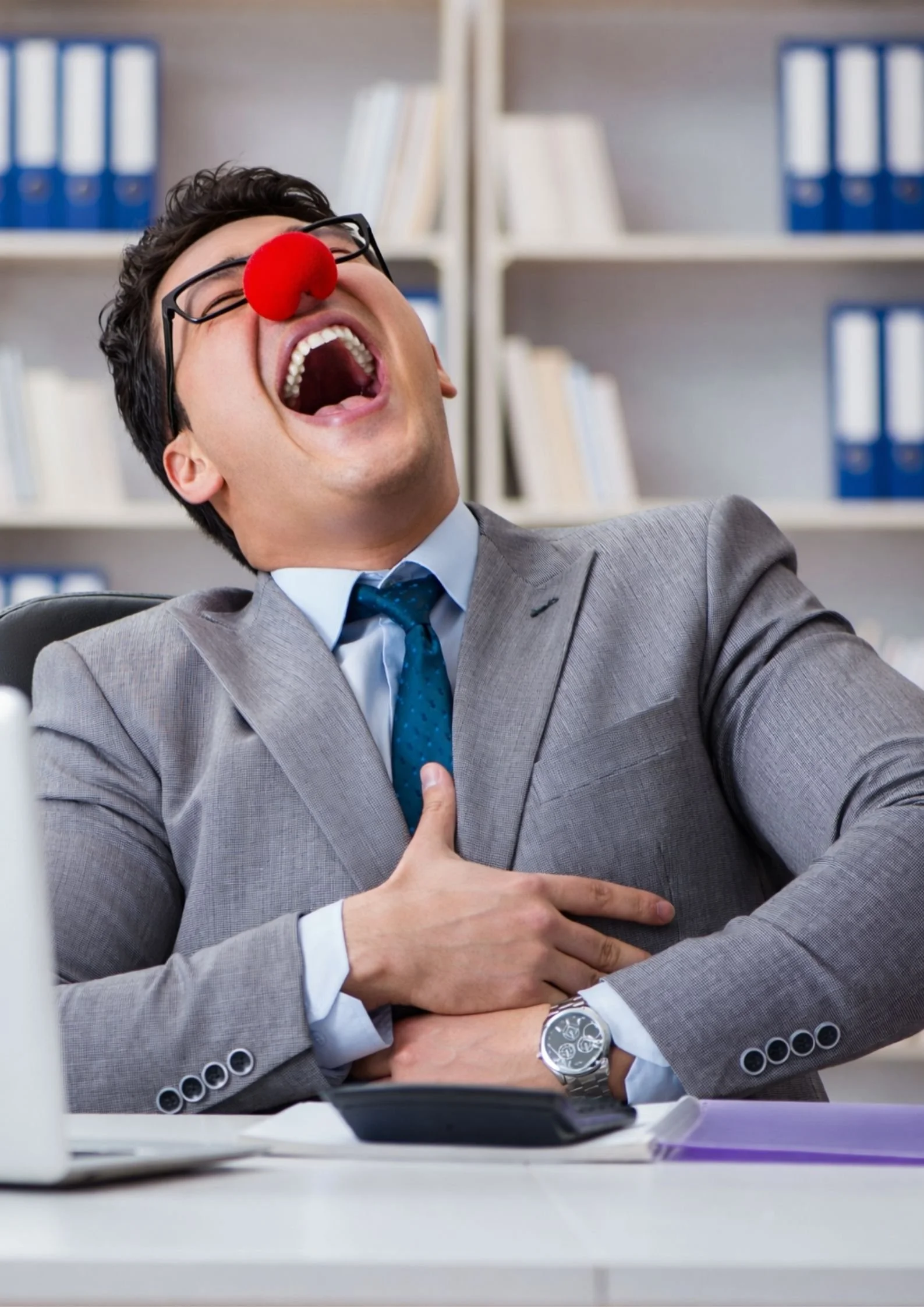 Man in a gray business suit with a red clown nose, glasses, and a wristwatch, laughing with his mouth wide open and hand on his chest, sitting at a desk with a computer and papers, with shelves of binders and books in the background.