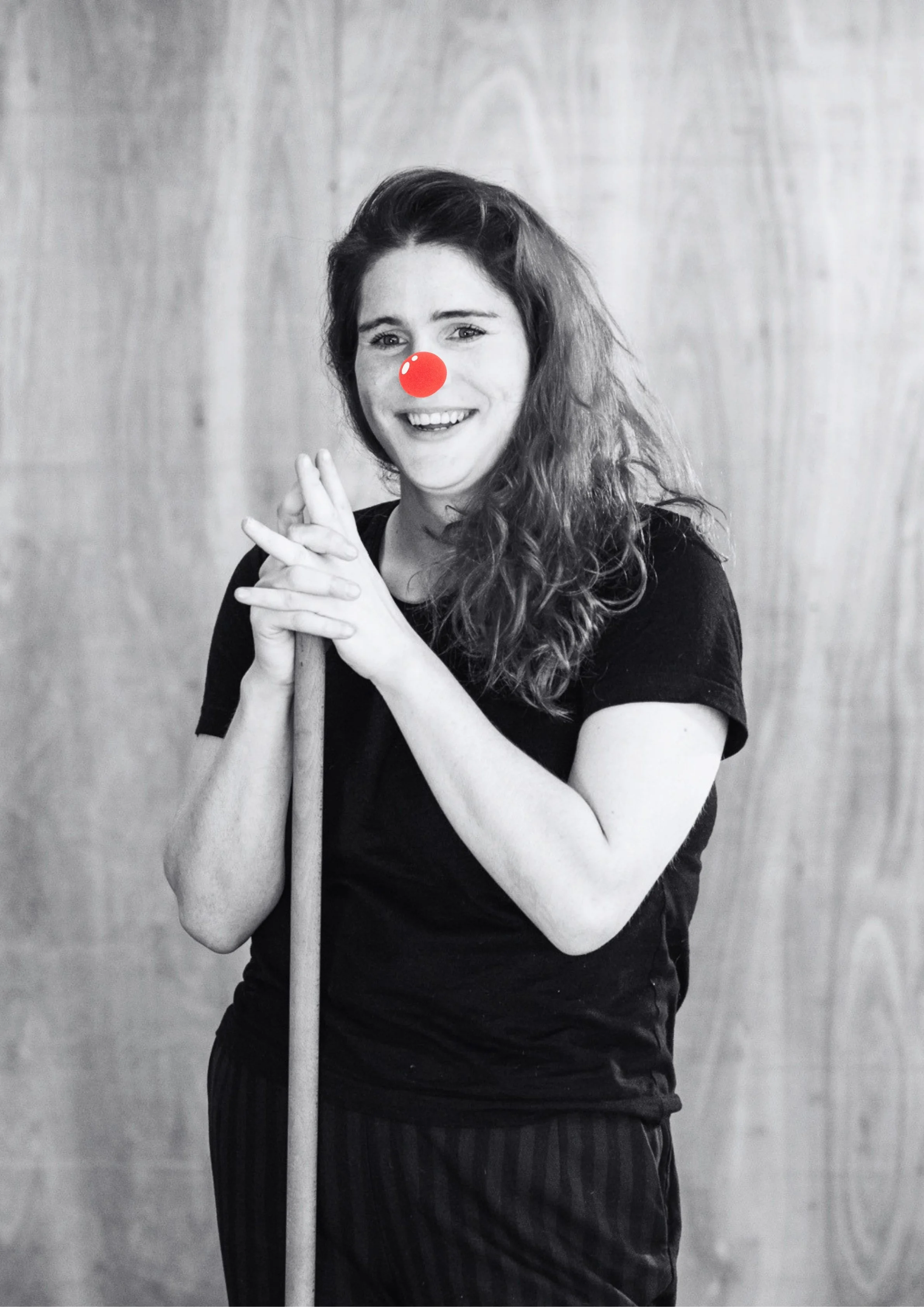 Black and white photo of a woman with wavy hair smiling and holding a cane, with a red clown nose added digitally.