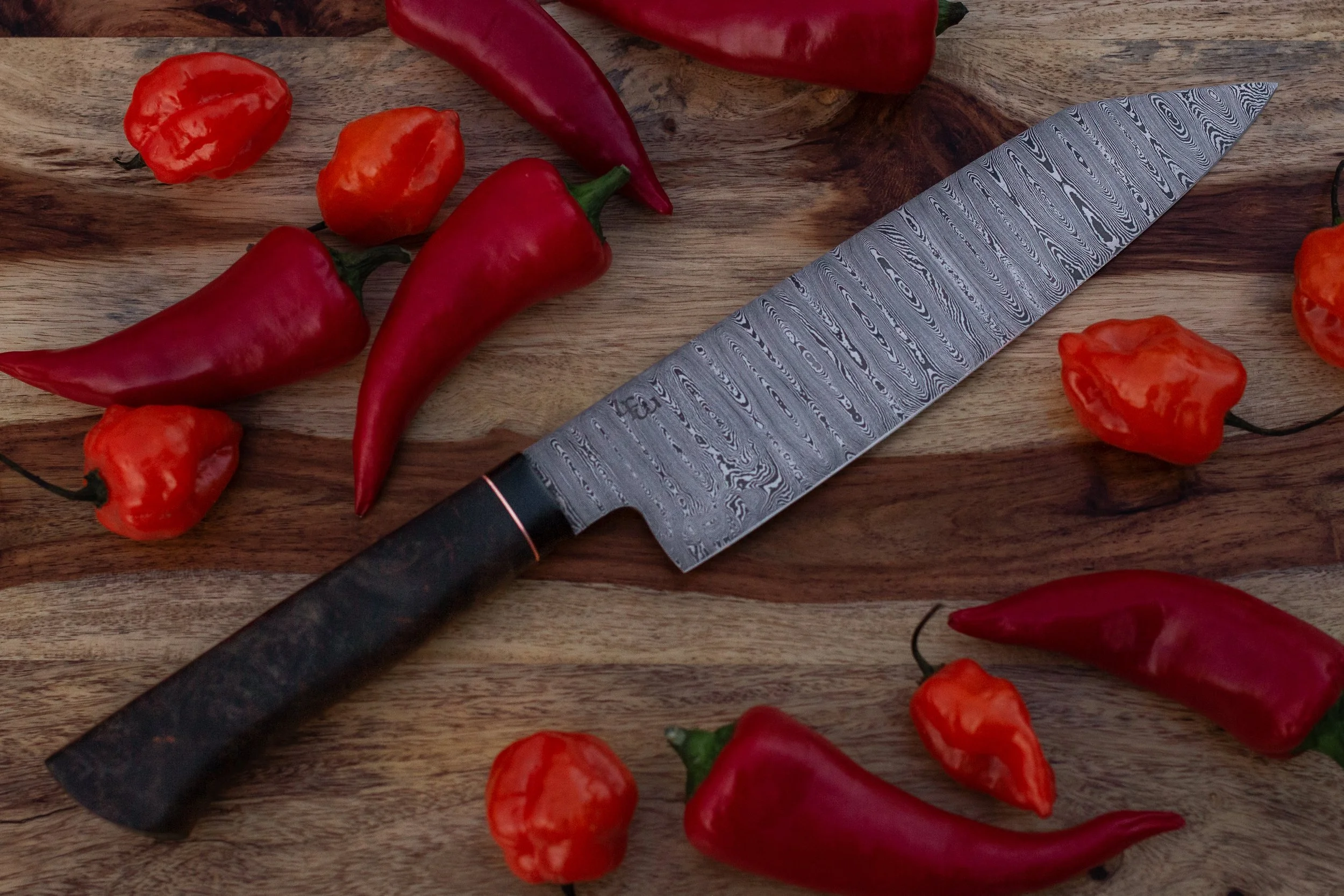Red chili peppers and orange habanero peppers on a wooden cutting board with a patterned chef's knife.