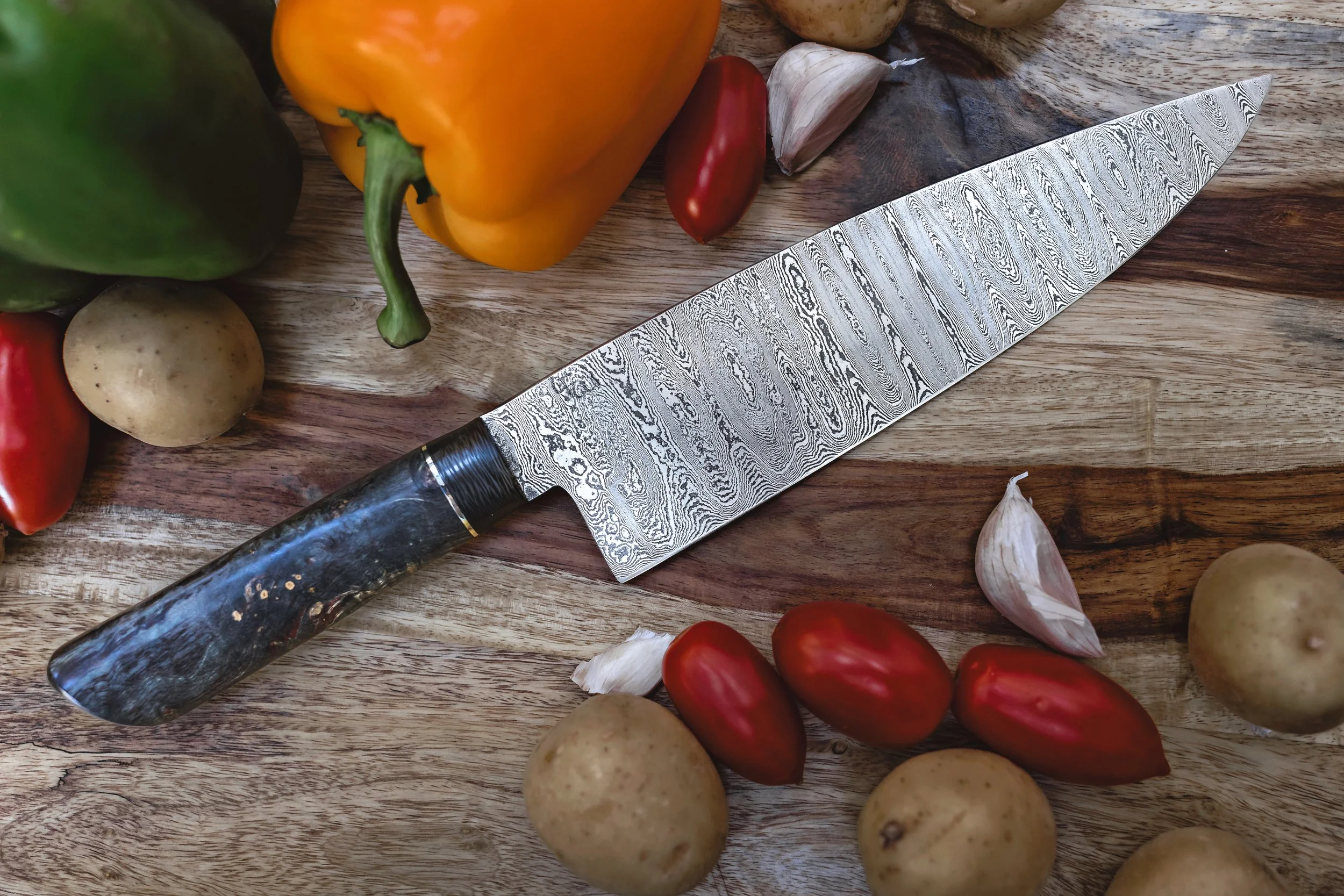 A patterned chef's knife with a dark marbled handle resting on a wooden surface surrounded by fresh vegetables including yellow, green, and red peppers, cherry tomatoes, garlic cloves, and potatoes.