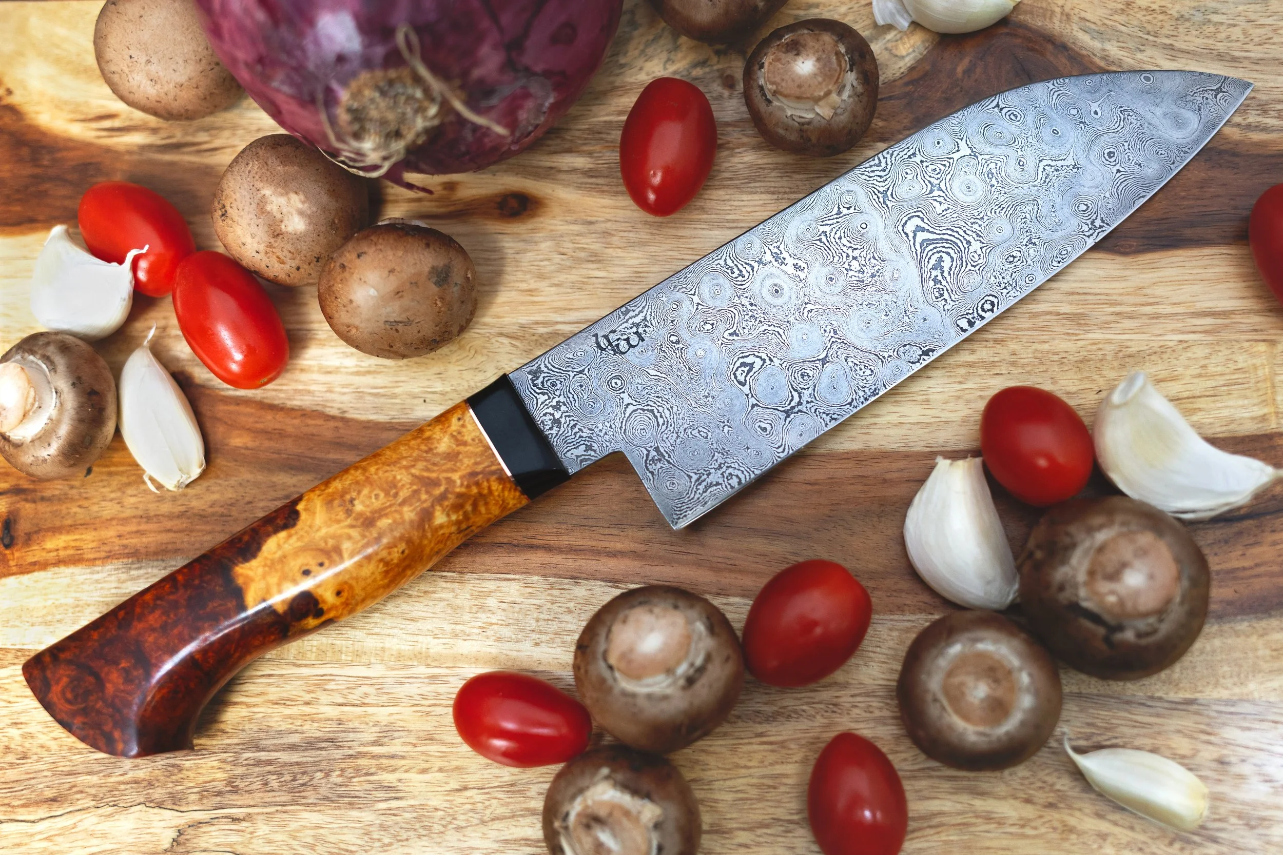 A kitchen knife lying on a wooden cutting board surrounded by garlic cloves, grape tomatoes, whole mushrooms, and a red onion.