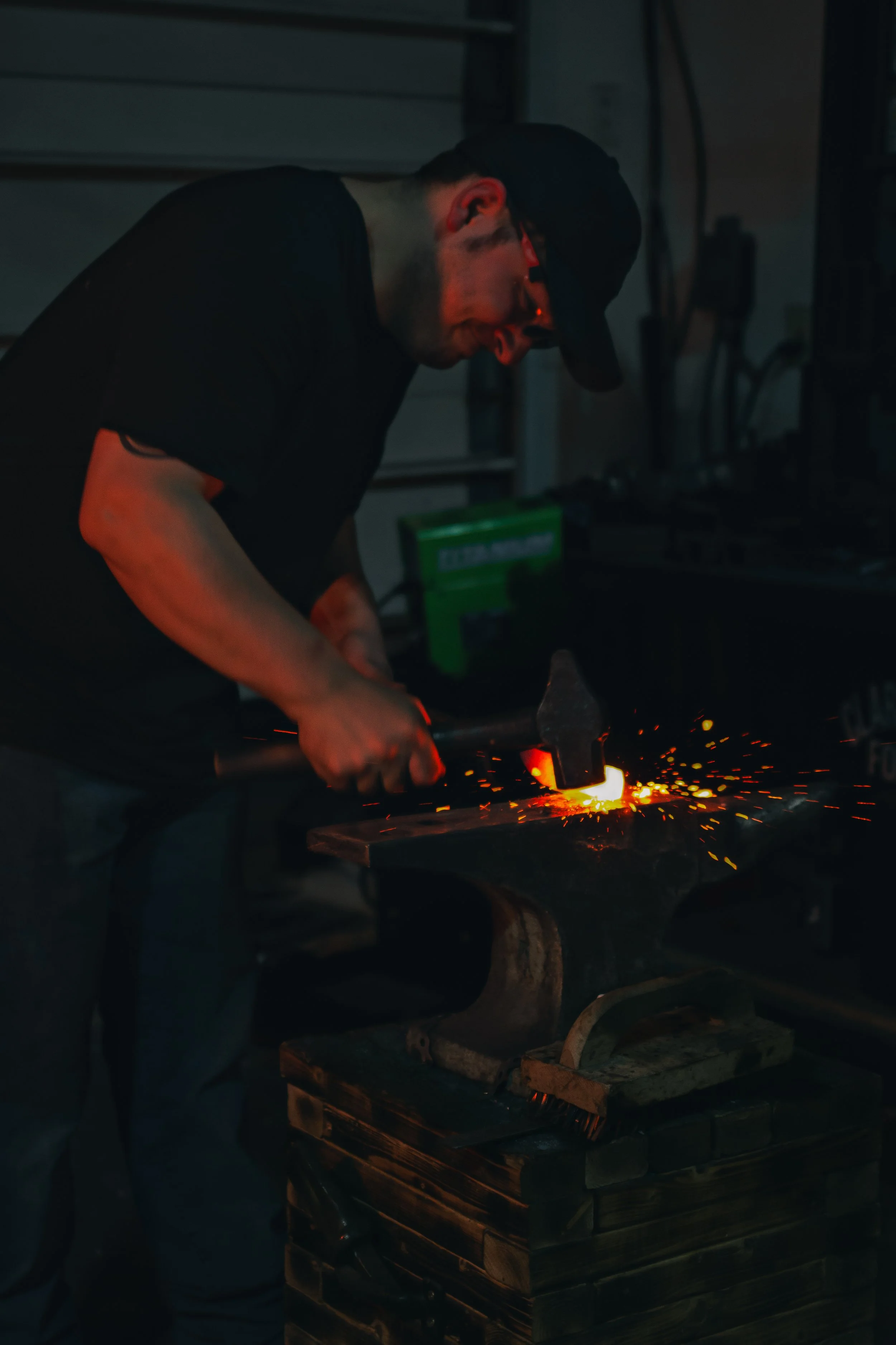 A man wearing a black cap and t-shirt is working on metal using a hammer and a fire while sparks fly in a dark workshop.