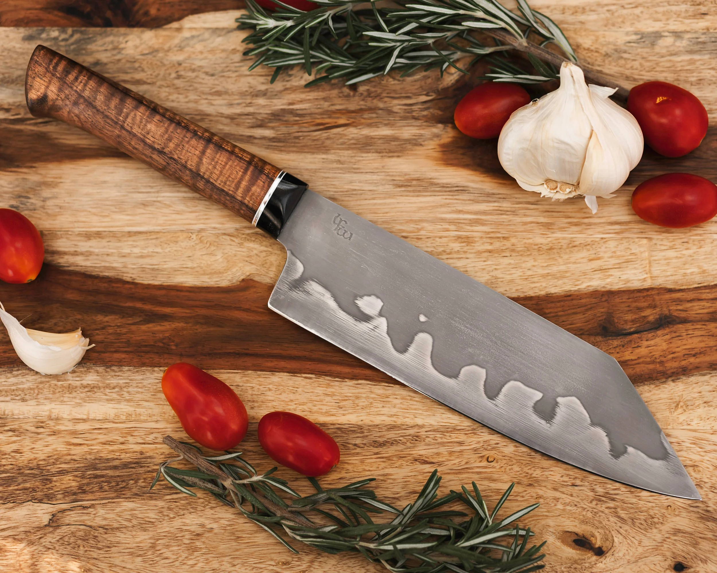A kitchen knife with a wooden handle resting on a wooden cutting board surrounded by cherry tomatoes, garlic cloves, and sprigs of rosemary.