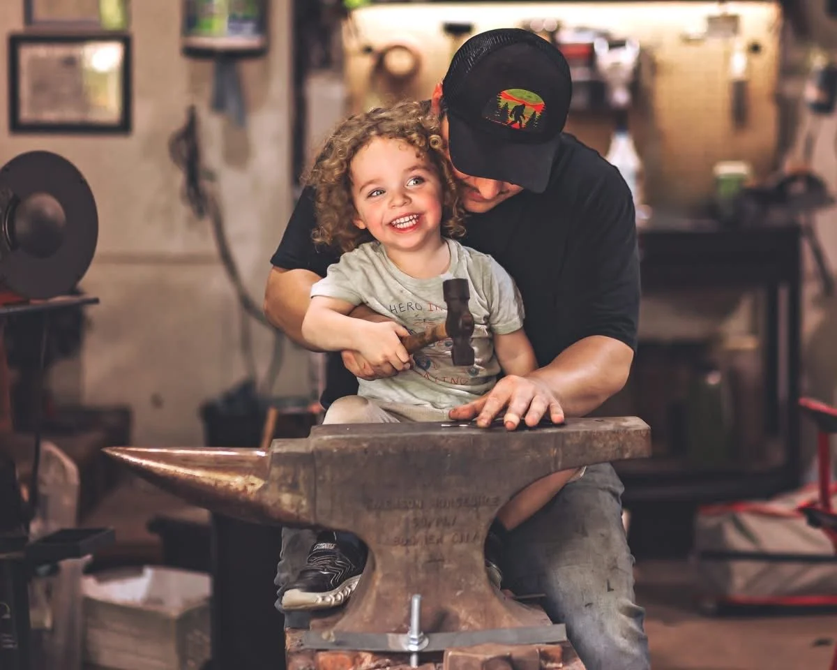 A child with curly hair holding a hammer while sitting on an anvil, with an adult helping and guiding them in a workshop or blacksmith's shop.