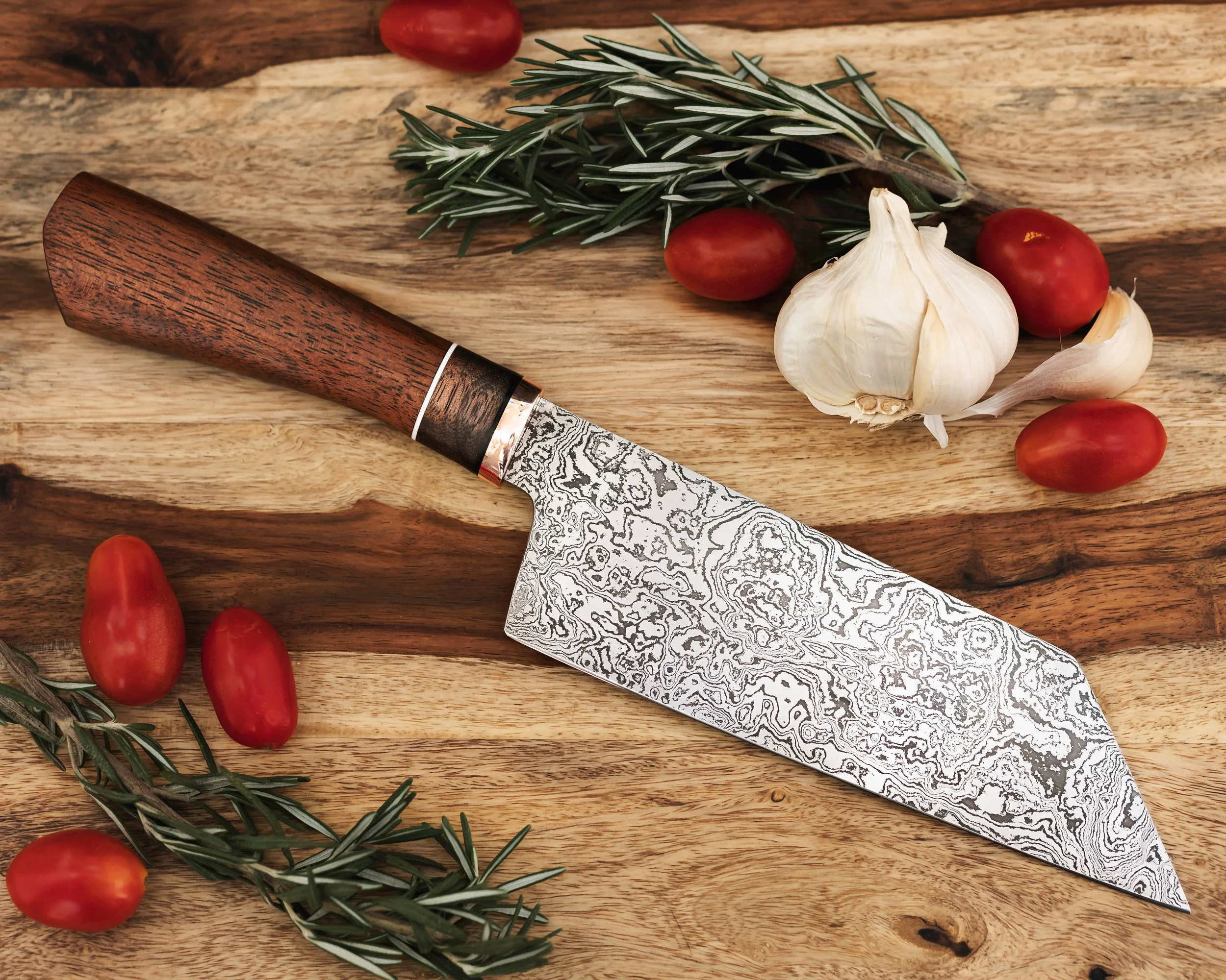 A patterned chef's knife with a wooden handle on a wooden cutting board, surrounded by cherry tomatoes, garlic bulb, and sprigs of rosemary.