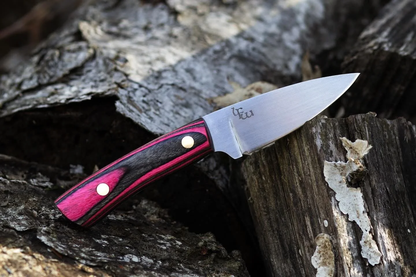 A pocket knife with a pink and black handle resting on a piece of chopped log.