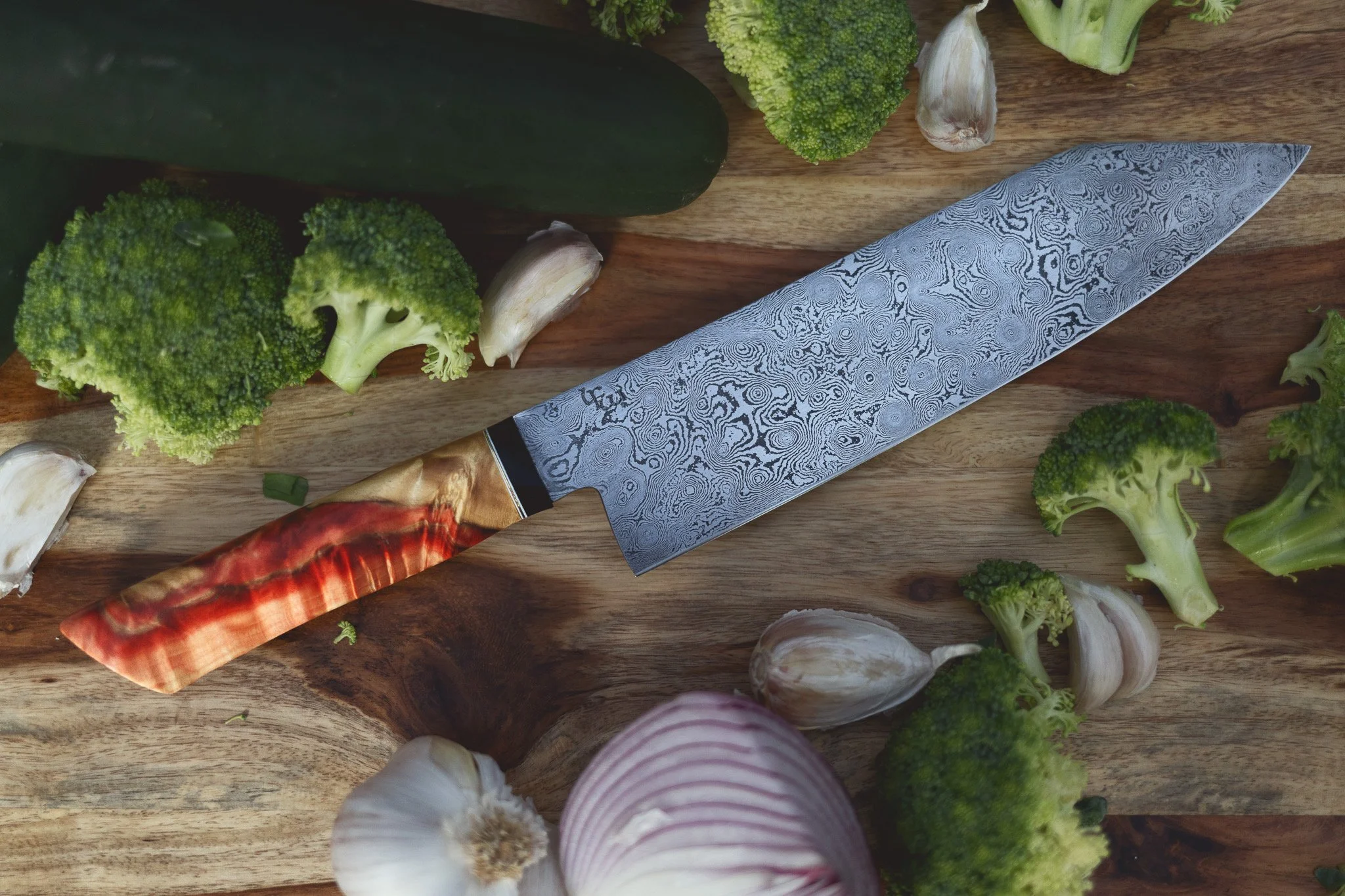 A Damascus steel chef's knife with a decorative handle placed on a wooden cutting board surrounded by broccoli, garlic, onion, cucumber, and other vegetables.