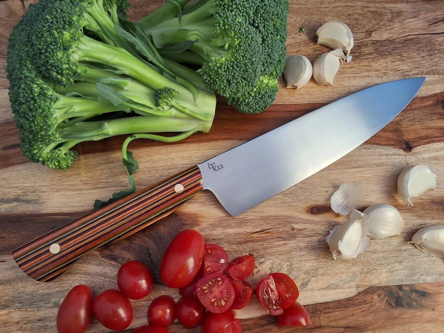 Fresh broccoli, garlic cloves, cherry tomatoes, and a kitchen knife on a wooden cutting board.