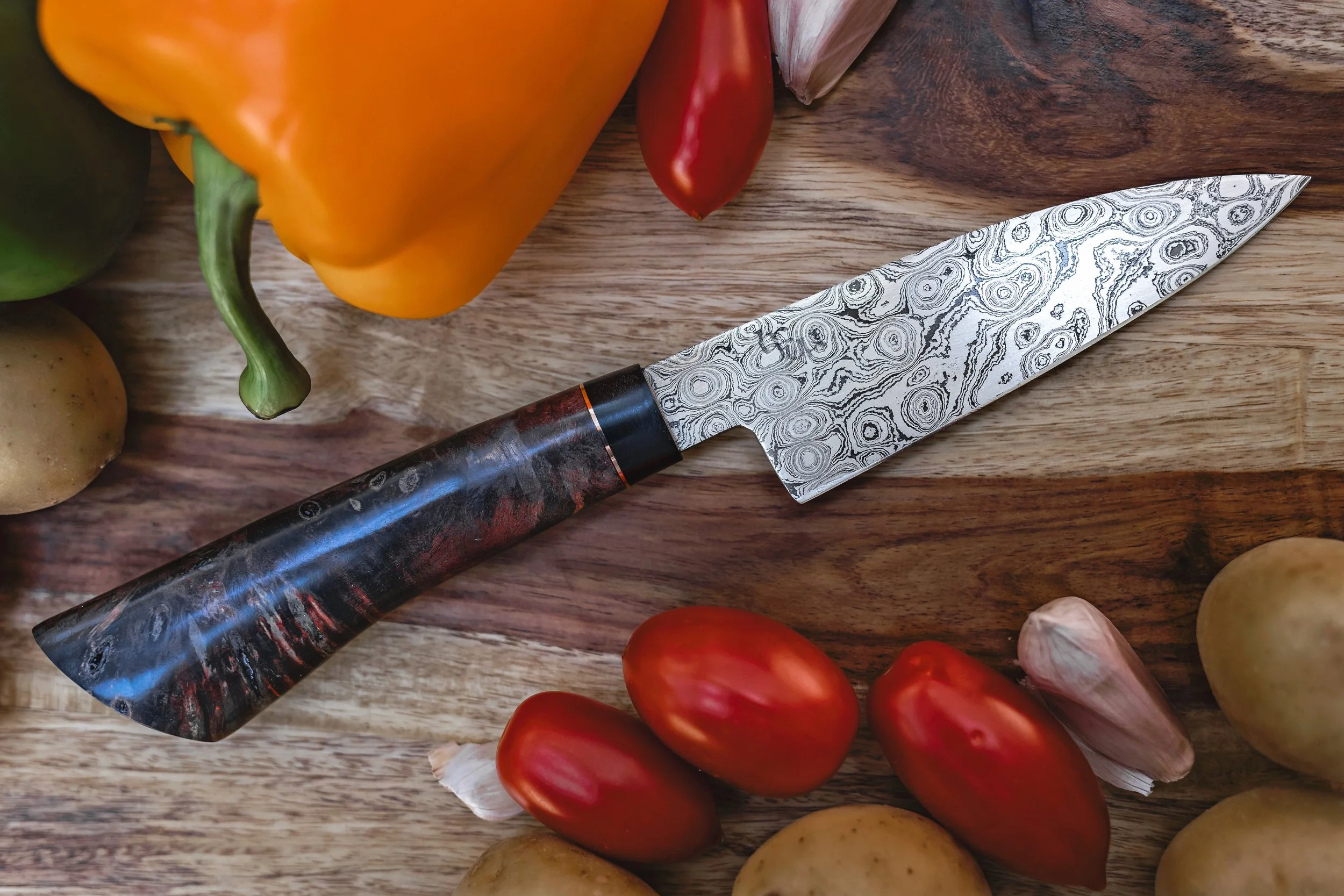 A patterned chef's knife with a marbled handle on a wooden cutting board surrounded by tomatoes, garlic, potatoes, and a yellow bell pepper.