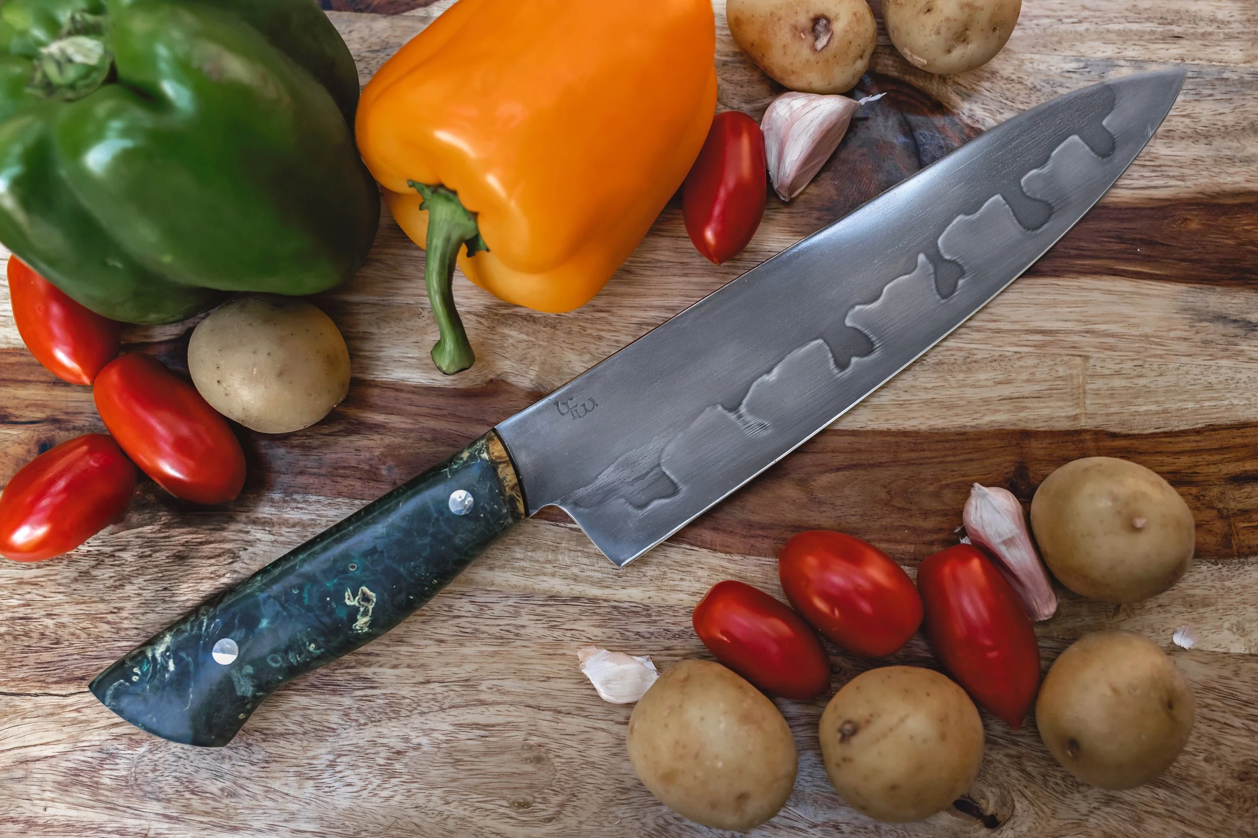 A kitchen countertop with a large chef's knife, colorful bell peppers, cherry tomatoes, potatoes, and garlic cloves.