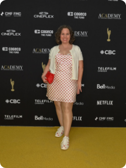 Woman standing on a yellow carpet in front of a black backdrop with various logos. She is wearing a white dress with red polka dots, a white cardigan, and white shoes. She is holding a red clutch bag and smiling at the camera.