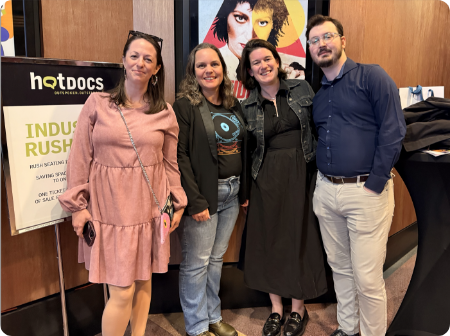 Four people standing together at an indoor event with a poster and artwork behind them.