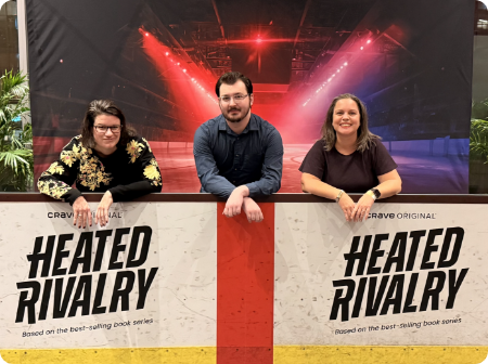 Lucy, Audrey and Liam - from Bizable - behind a table with promotional signs for 'Heated Rivalry' at a Canadian Television event.