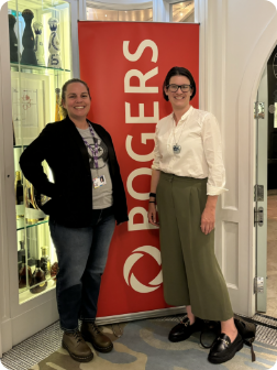 Lucy and Audrey - co presidents of Bizable standing in front of a red and white Rogers banner inside a building at a Canadian Film and Television event, with shelves of decorative items and a glass door in the background.
