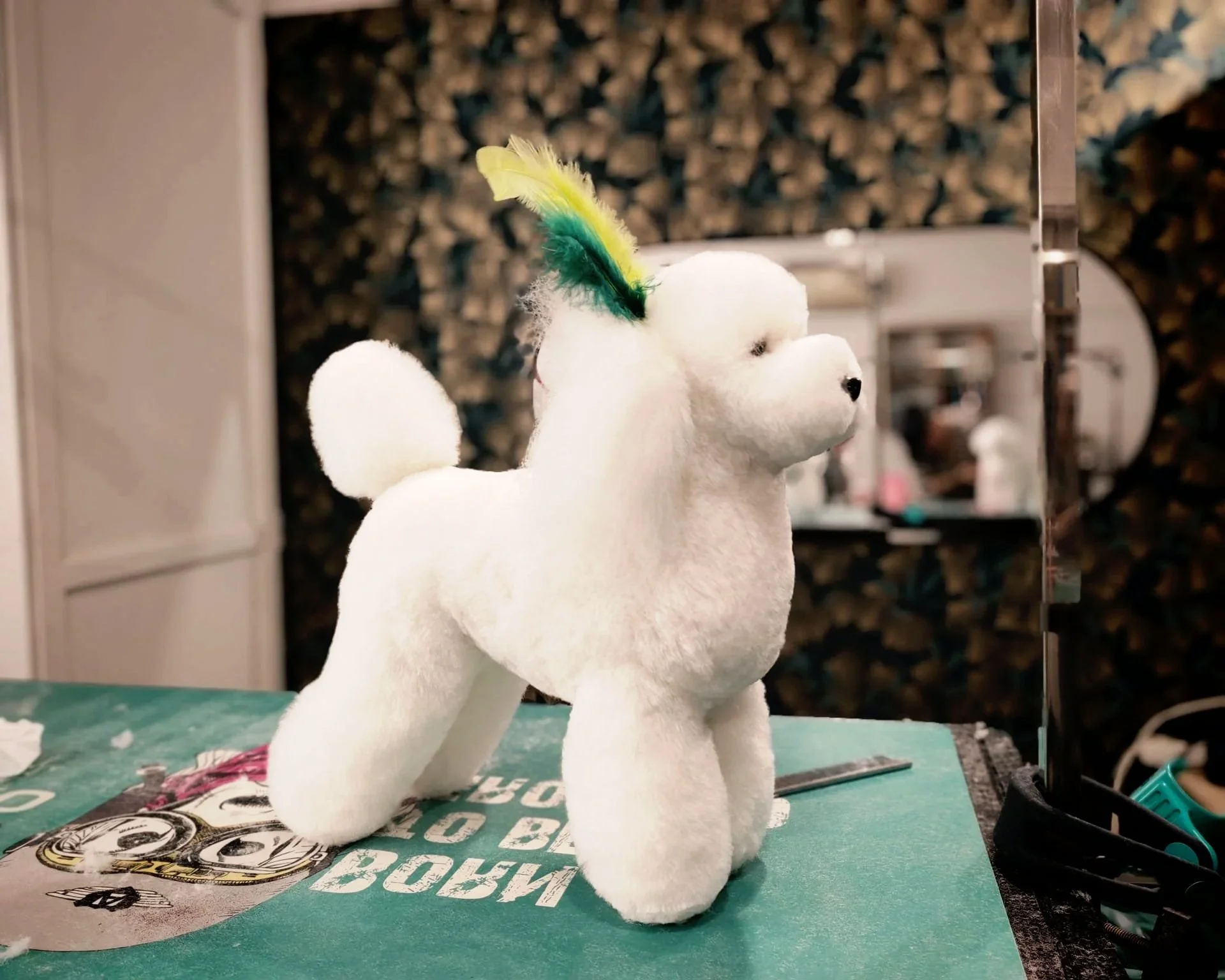 A plush toy dog with a feathered crest on its head, standing on a work surface in a workshop or grooming area, with a background of a textured wall and grooming tools.
