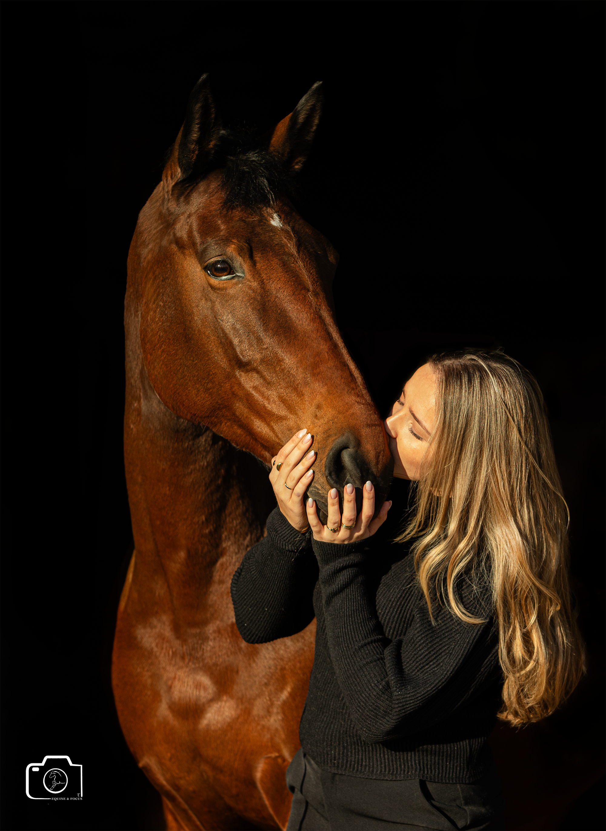 A woman with long blonde hair embracing and kissing a brown horse with a black mane against a black background.