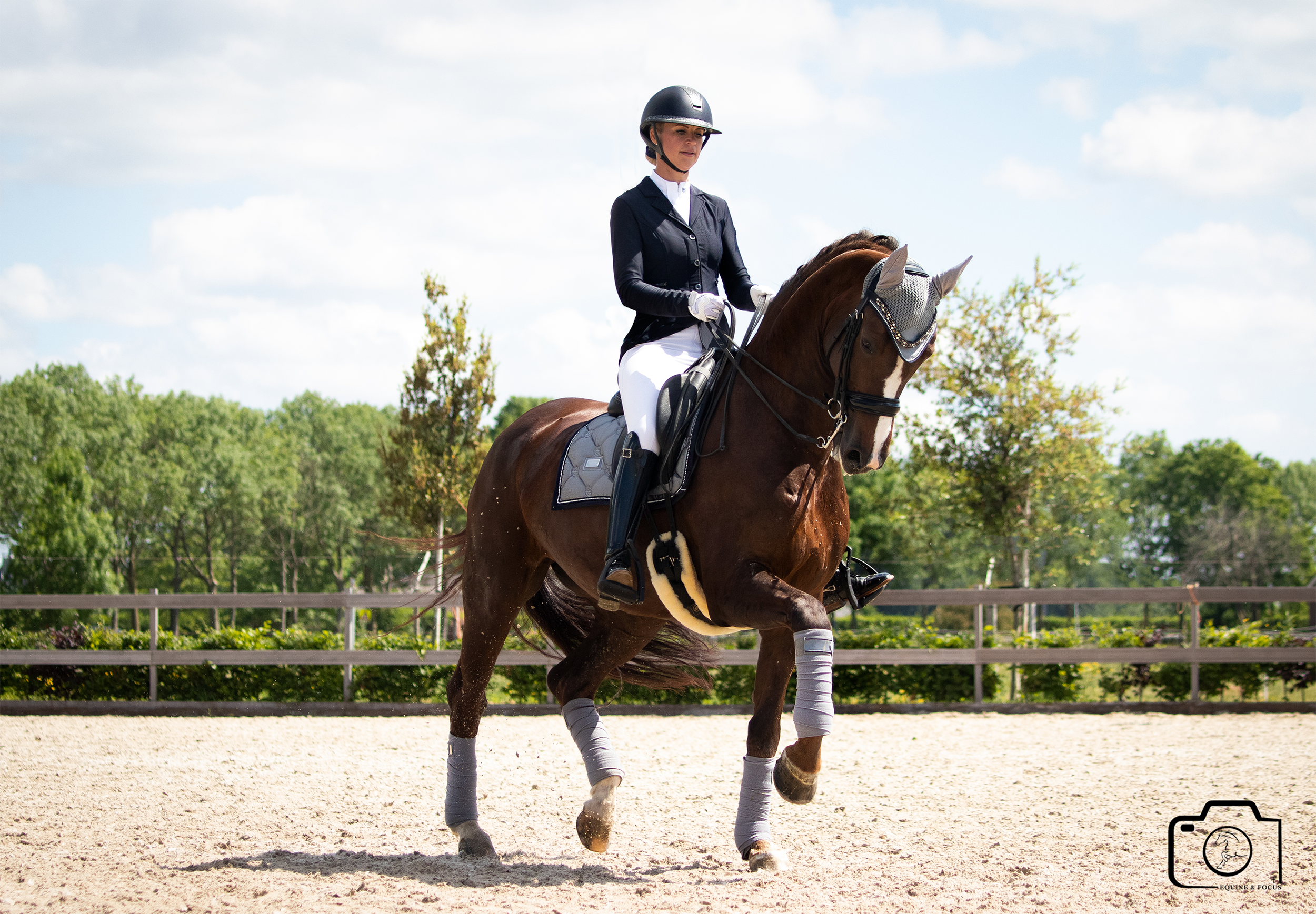 A woman riding a brown horse on a sandy outdoor riding arena. She is wearing a black helmet, black riding jacket, white breeches, and black riding boots, with some paddings on the horse's legs. The background features trees and a partly cloudy sky.