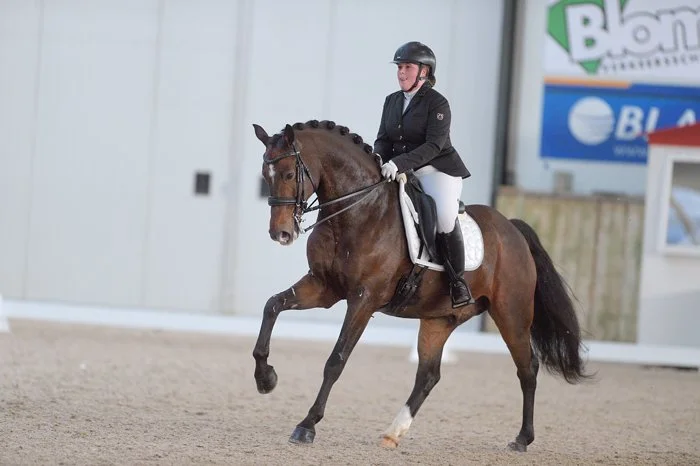 A female equestrian riding a brown horse with black mane and tail in an indoor arena, wearing a helmet, jacket, white pants, and gloves.