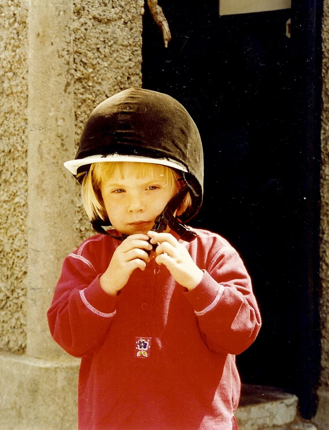 A young child with blonde hair wearing a black helmet and a red jacket, holding the helmet's chin strap with both hands, standing outdoors in front of a beige stone wall and a black door.