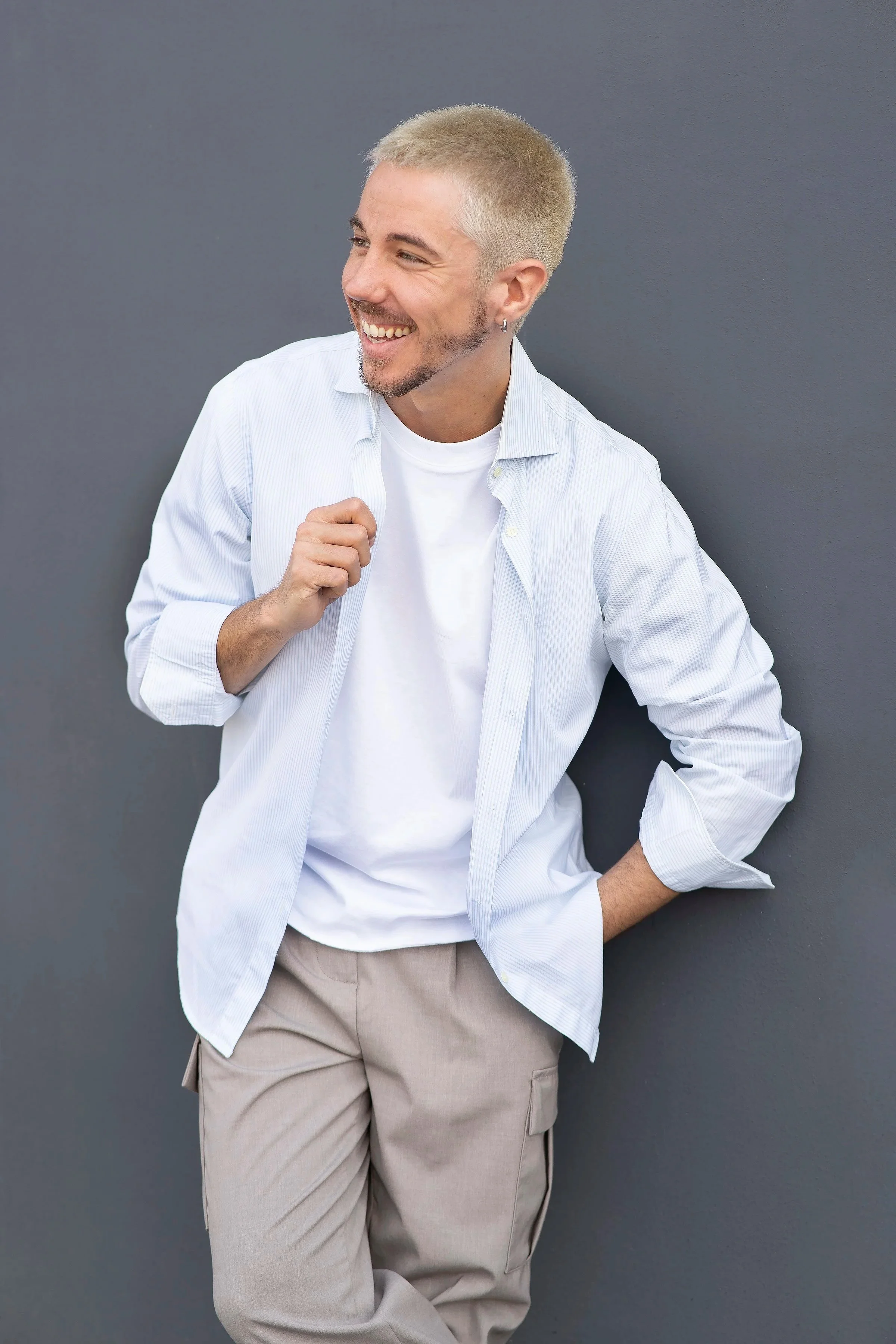 Uomo che sorride, indossa una camicia bianca e pantaloni beige, appoggiato a un muro grigio.