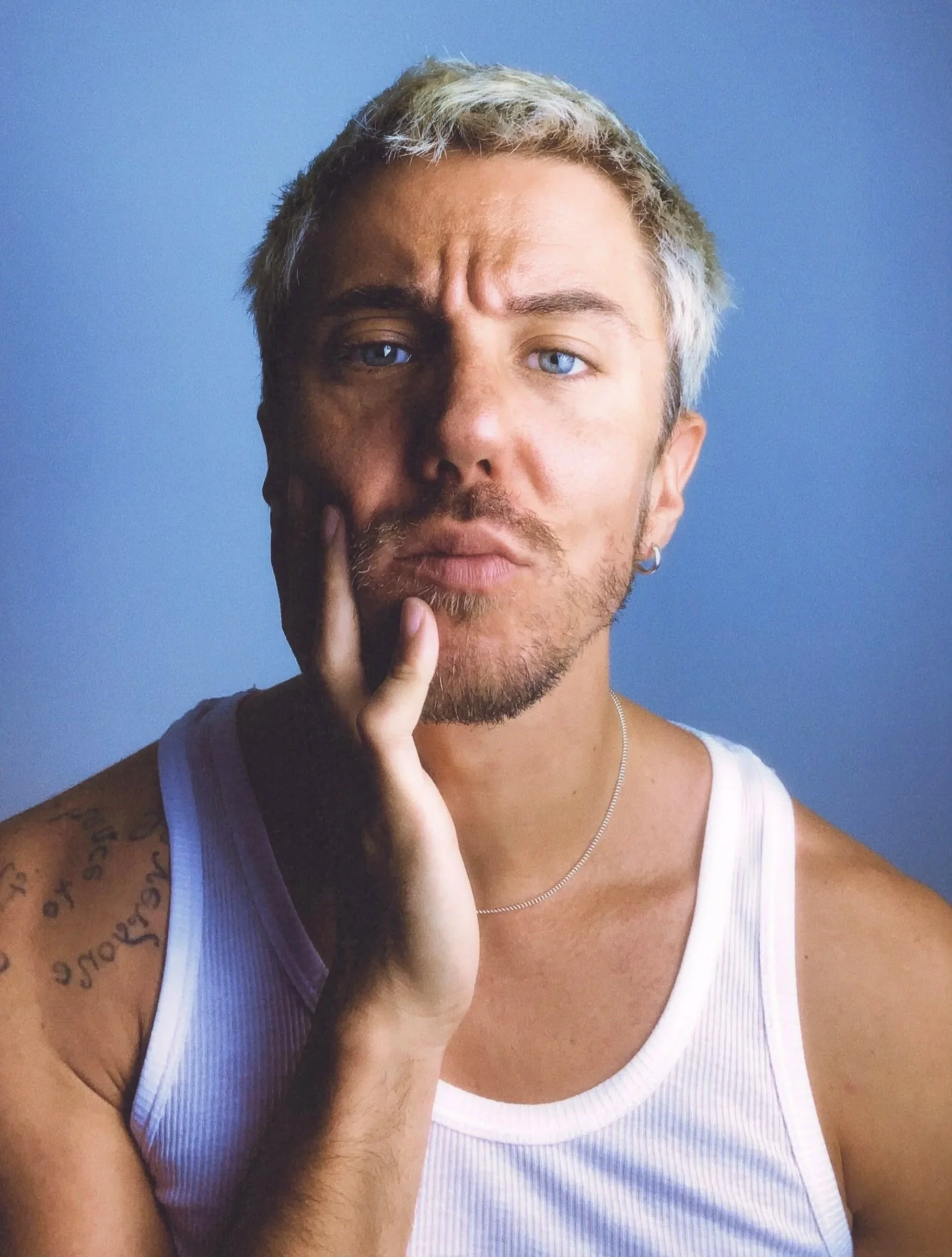 Ritratto di un giovane uomo con capelli biondi e occhi blu, con un orecchino, che indossa una canotta bianca e una collana, con uno sfondo blu.