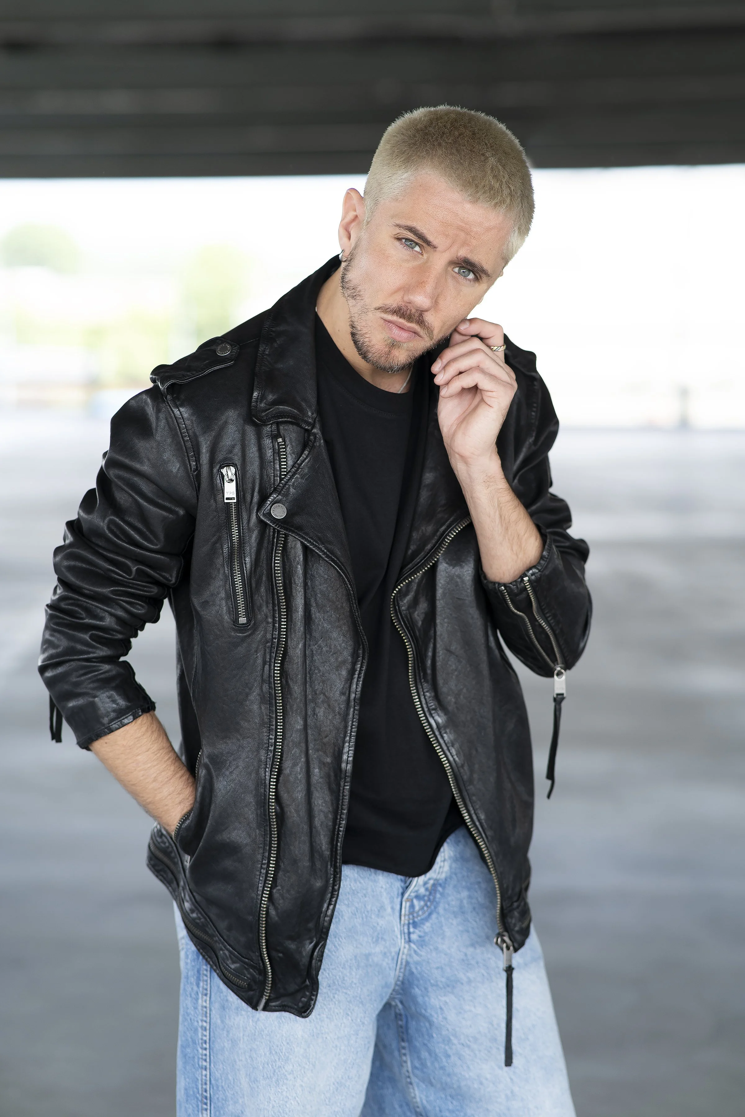 Uomo con capelli corti e biondi, indossa una giacca di pelle nera e una maglietta nera, in un ambiente aperto con un soffitto sotto un ponte o un'area coperta.