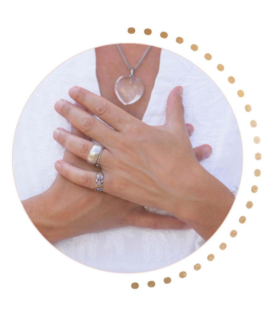 Close-up of a person's hands placed over their heart, wearing rings and a necklace with a heart-shaped pendant, against a white fabric background.