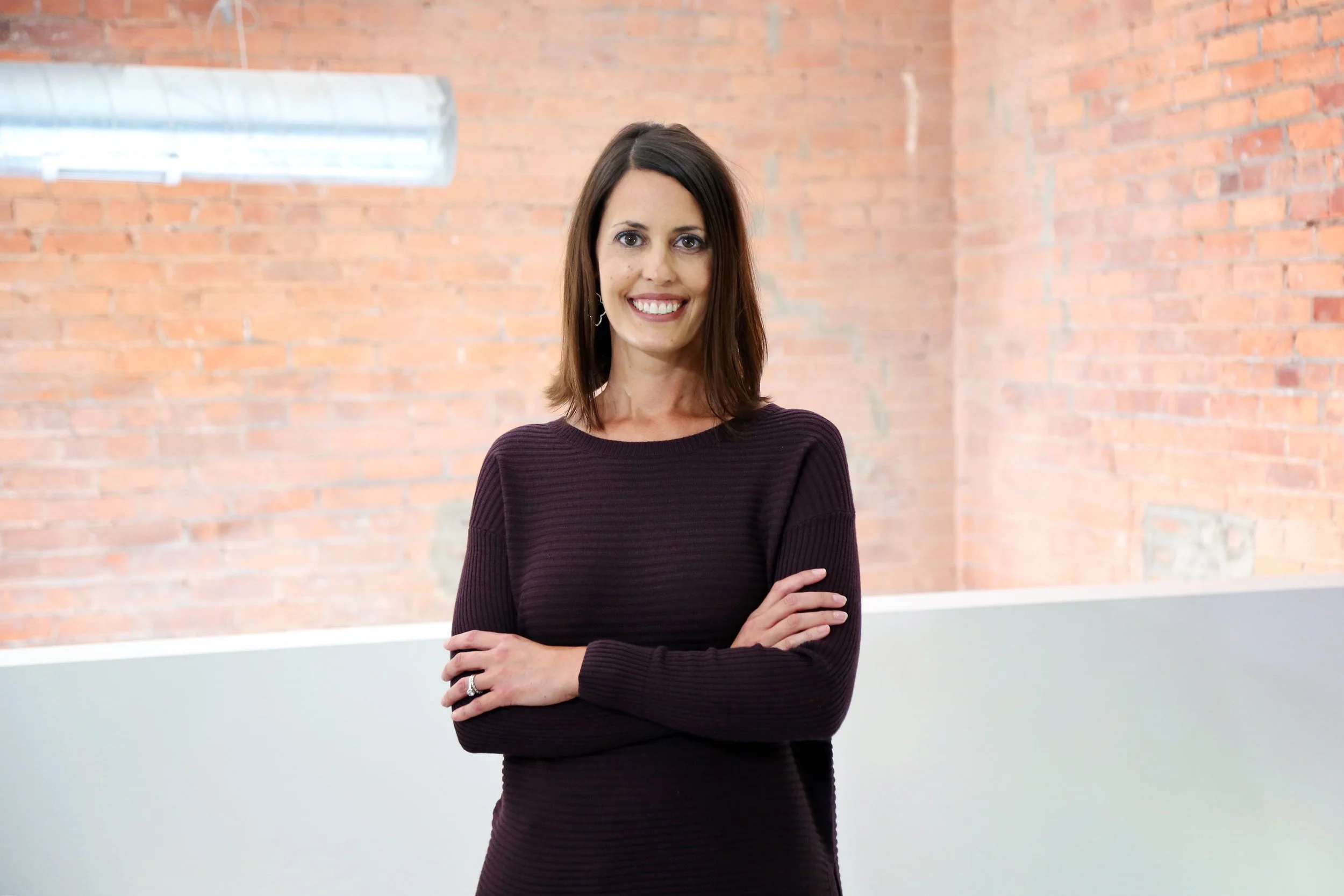 A woman with shoulder-length brown hair, wearing a dark long-sleeve top, smiling and crossing her arms, standing in front of a brick wall with a window and fluorescent light.