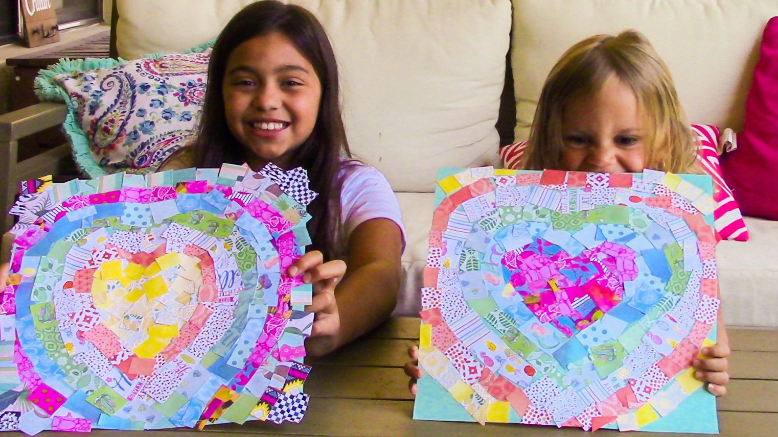 Two young girls sitting at a table holding colorful heart-shaped collage art pieces made from various patterned paper scraps.