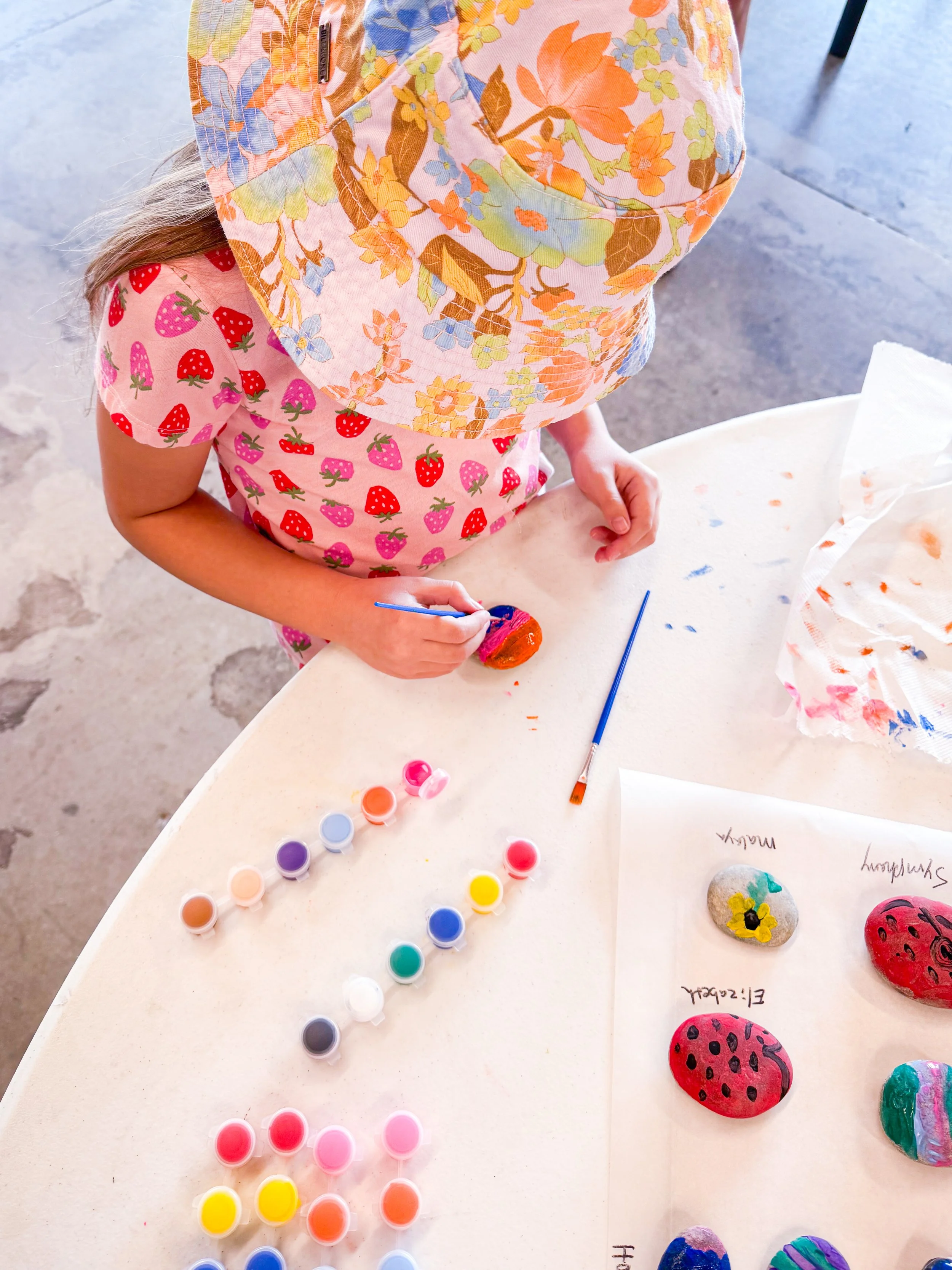 A child in a pink strawberry-patterned shirt and floral hat painting rocks with bright colors at a table, with paint pots and paper on the surface.