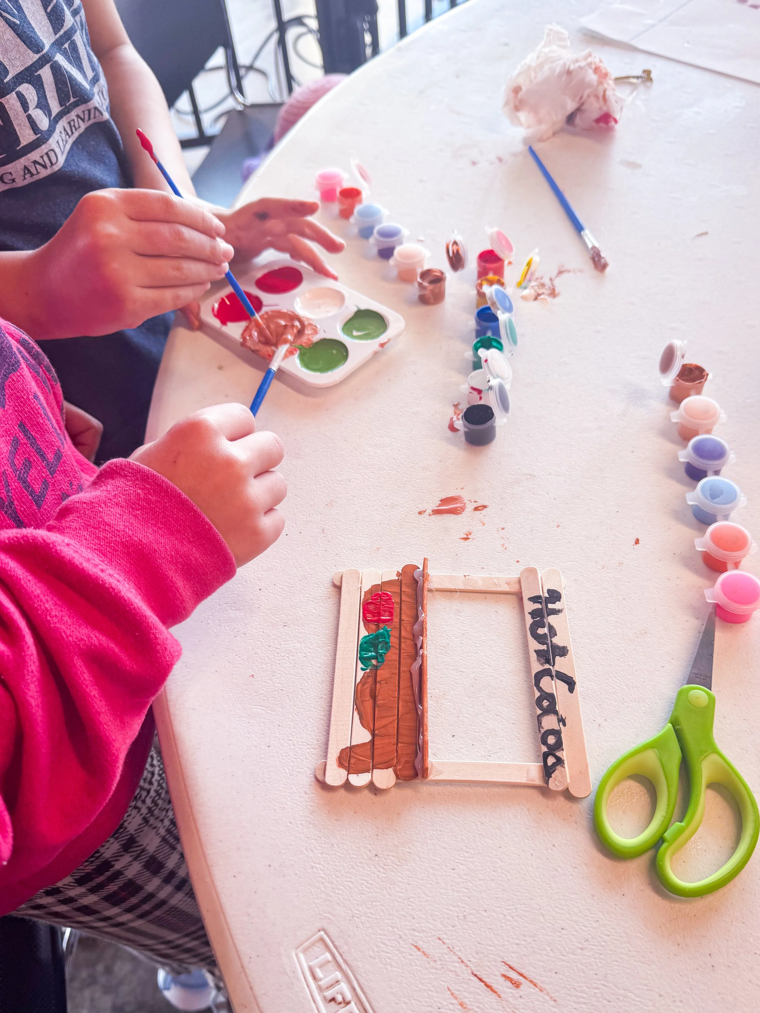 Children painting and decorating small wooden frames with brown and red paint at a table, with craft supplies like paint pots, scissors, and a paintbrush scattered on the table.