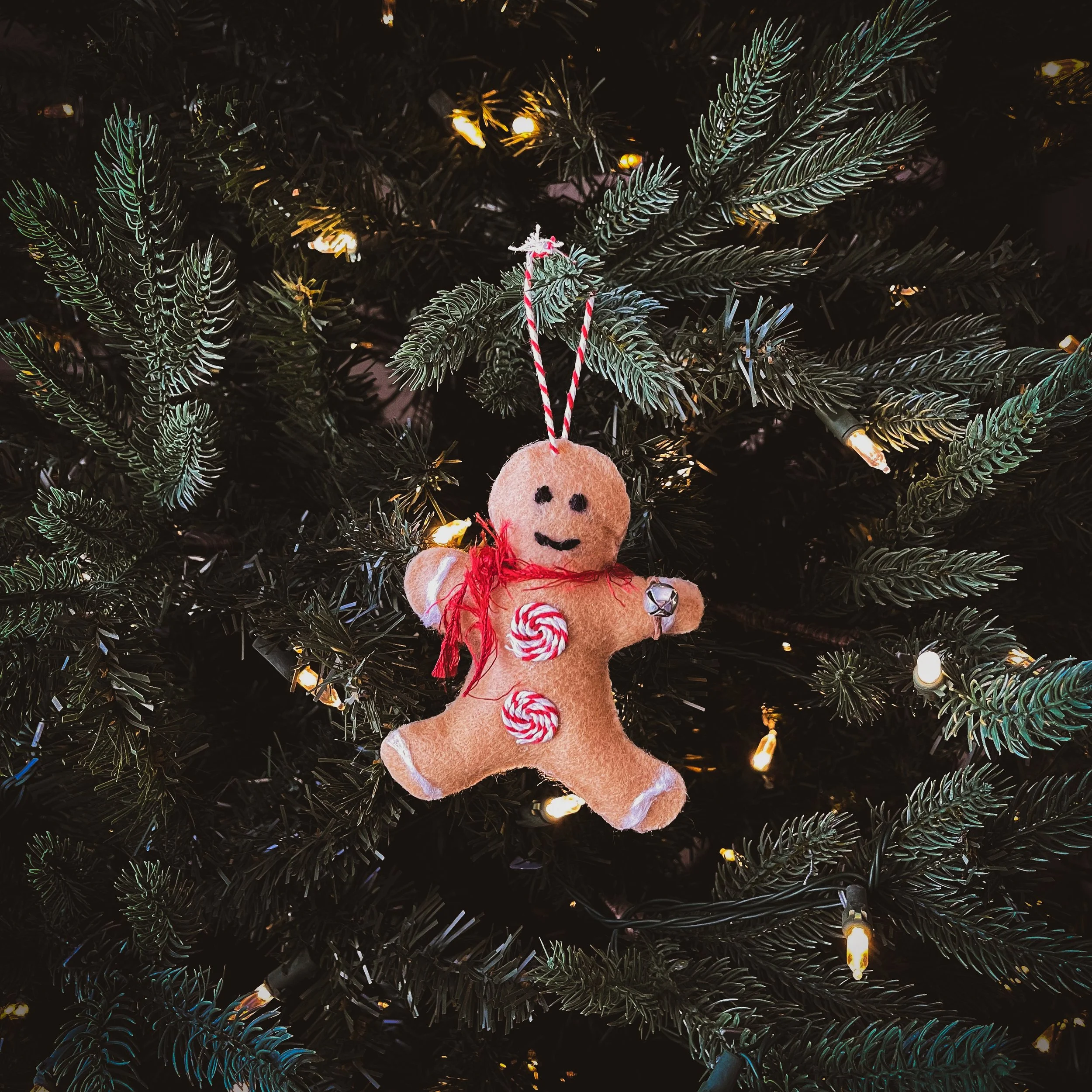 A gingerbread man Christmas tree ornament with a red bow, peppermint candies, a jingle bell, and a candy cane attached to its body, hanging on a decorated Christmas tree with lights.